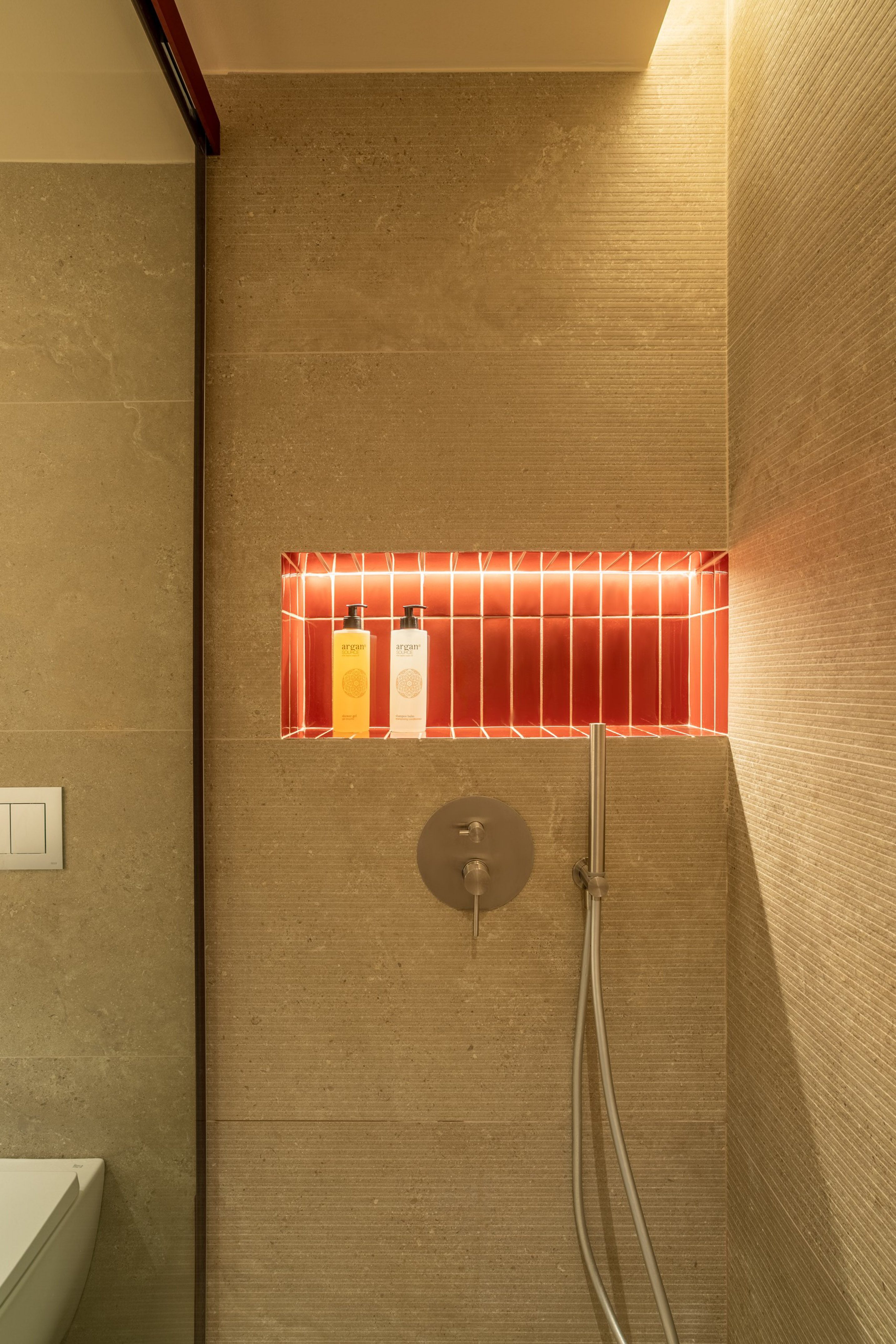 Modern shower with backlit red tile niche, argan products, and textured beige walls.