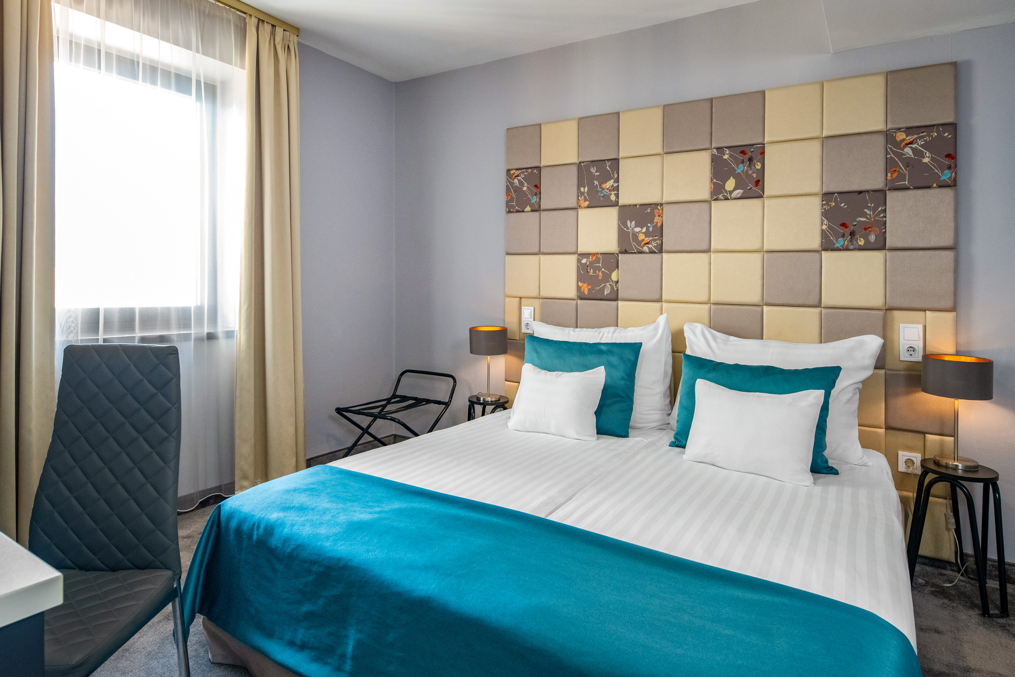 Modern hotel room with teal bedding, patterned headboard, and sunny window.