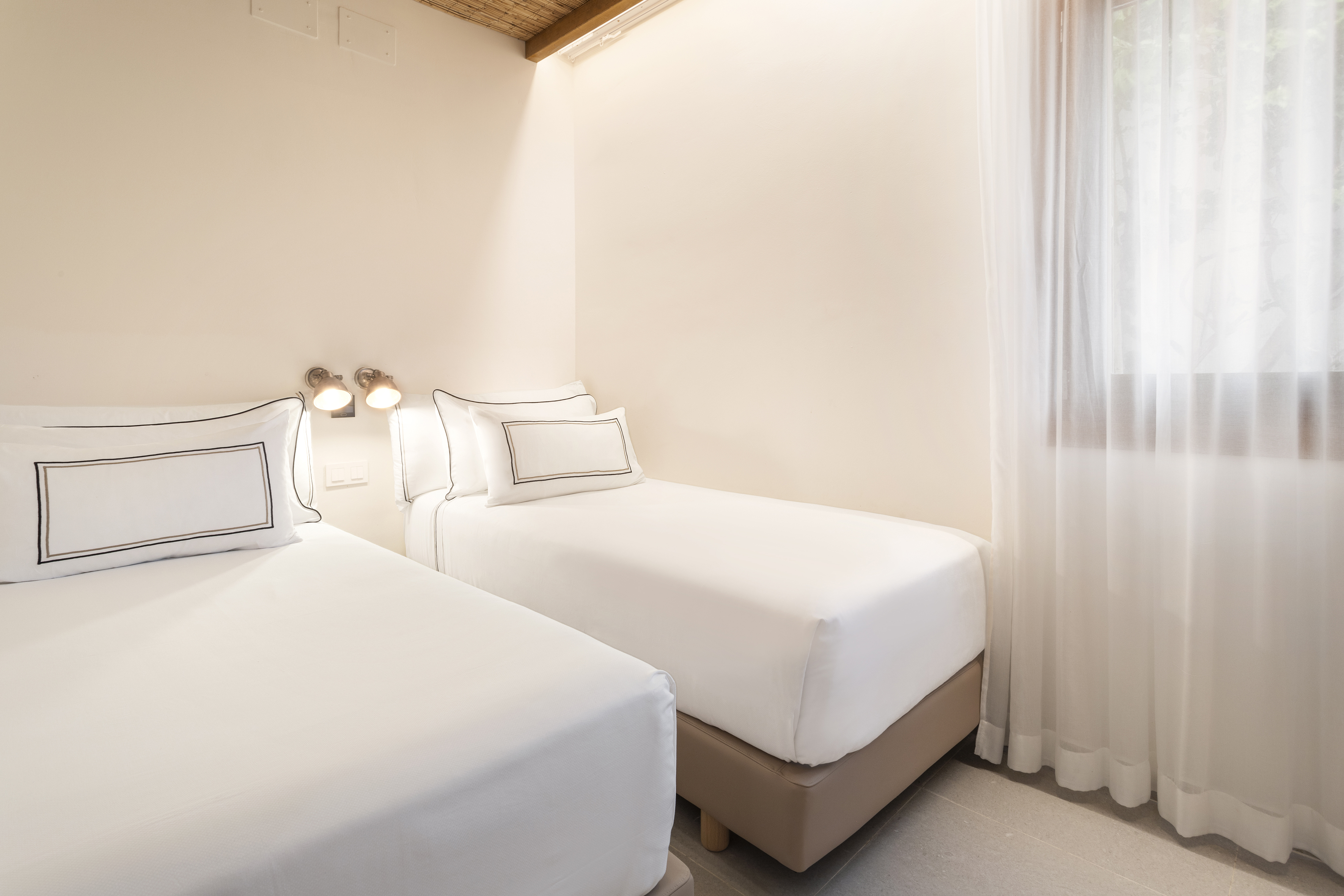 Modern bedroom with two twin beds, white linens, and sheer curtains.