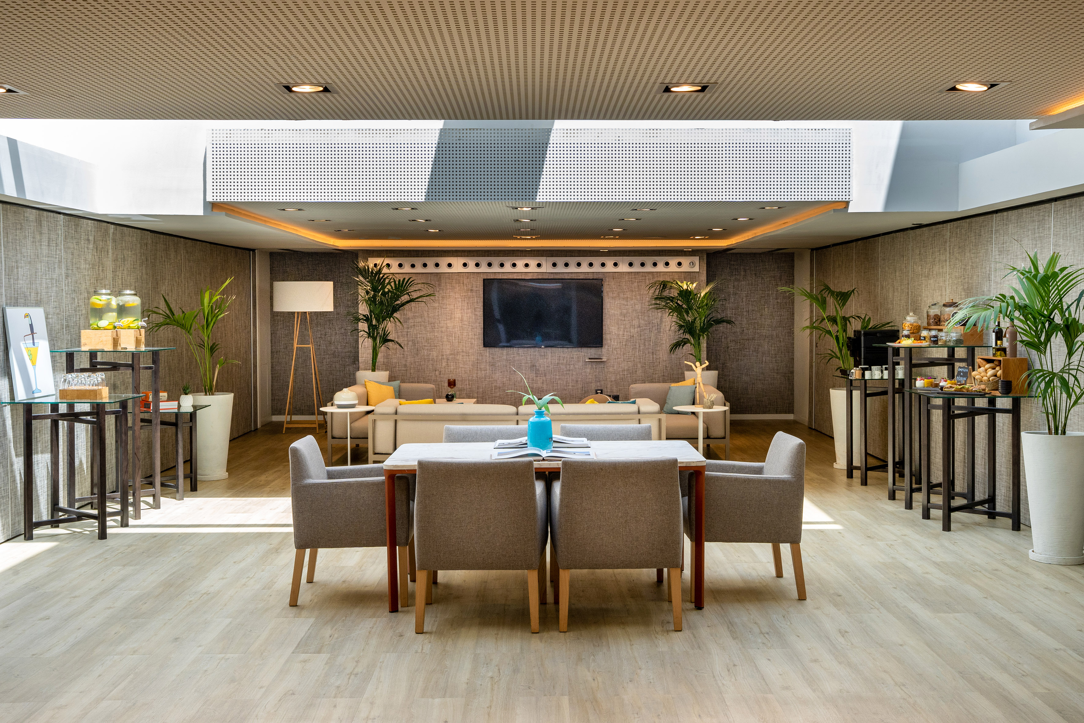 Modern hotel lounge with dining table, sofas, TV, and refreshment stations.