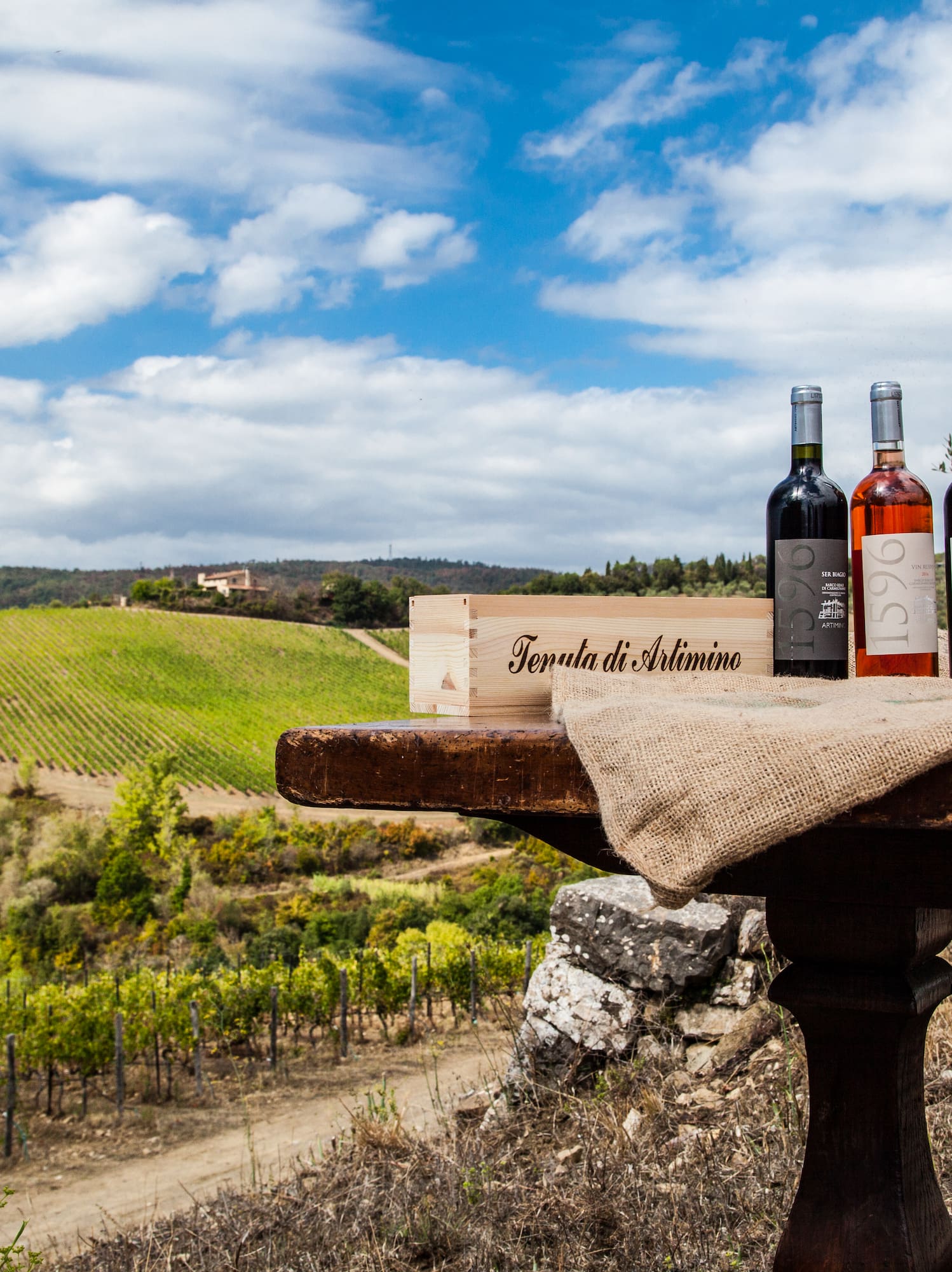 Artimino wine bottles, glass, crate on table with scenic Tuscan vineyard.
