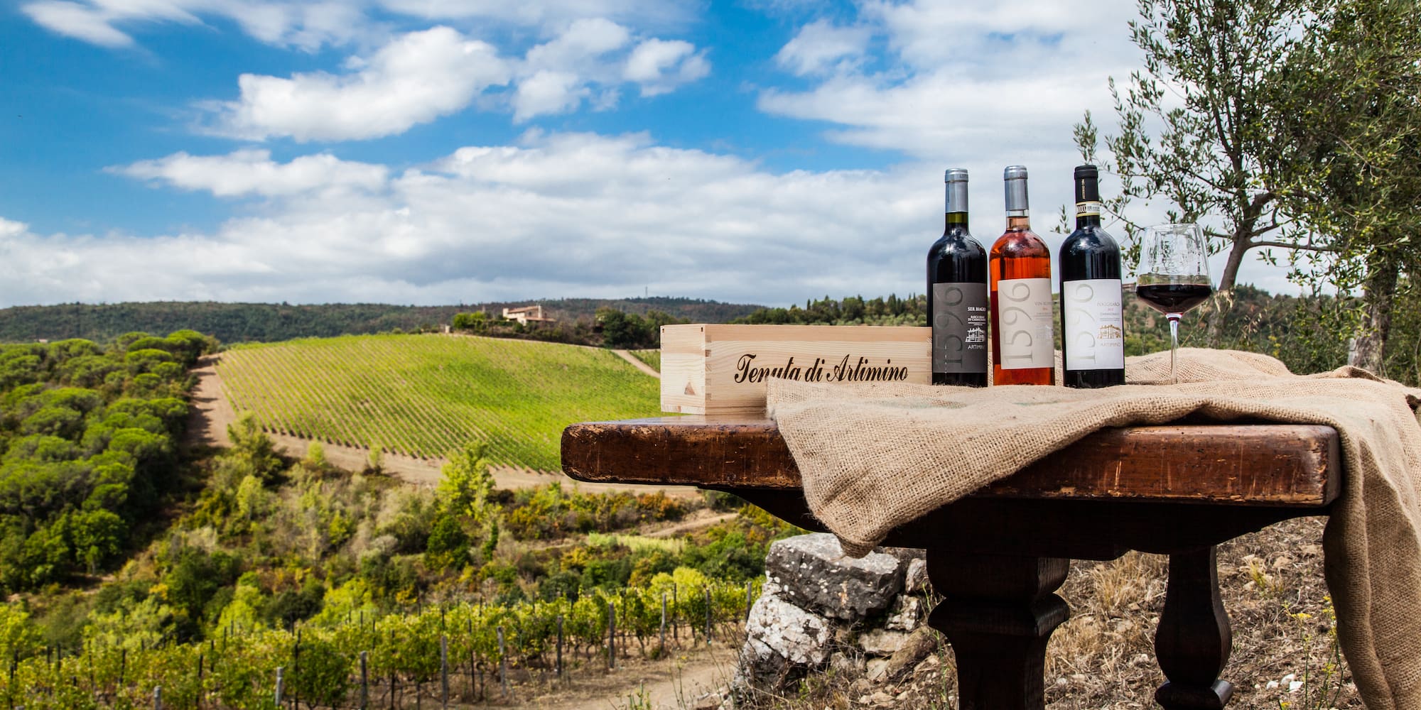 Artimino wine bottles, glass, crate on table with scenic Tuscan vineyard.