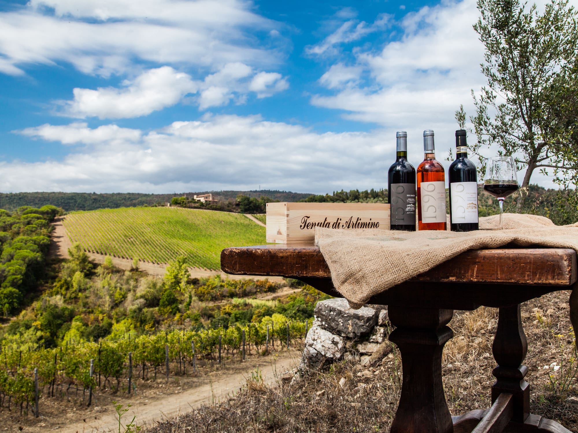 Artimino wine bottles, glass, crate on table with scenic Tuscan vineyard.
