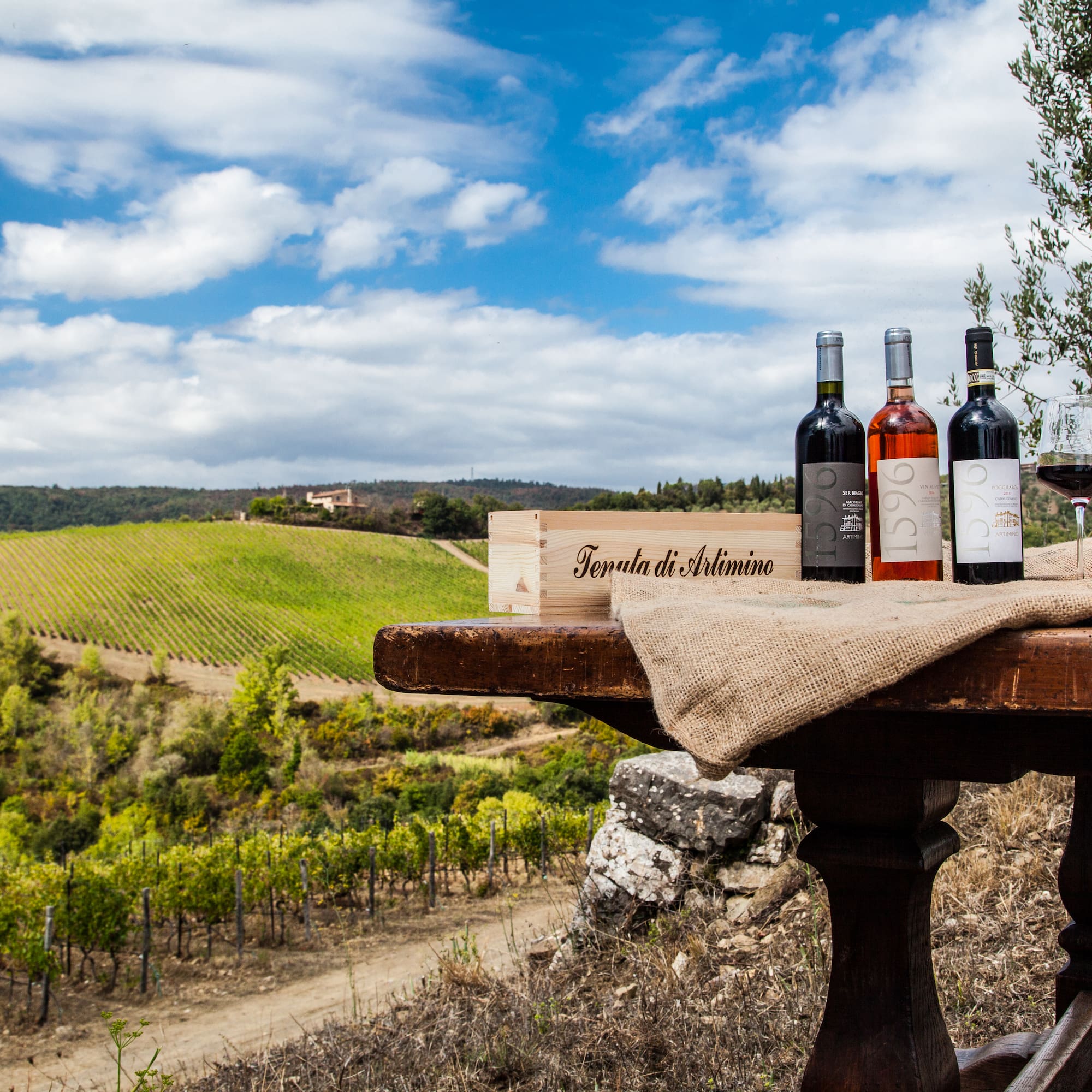 Artimino wine bottles, glass, crate on table with scenic Tuscan vineyard.