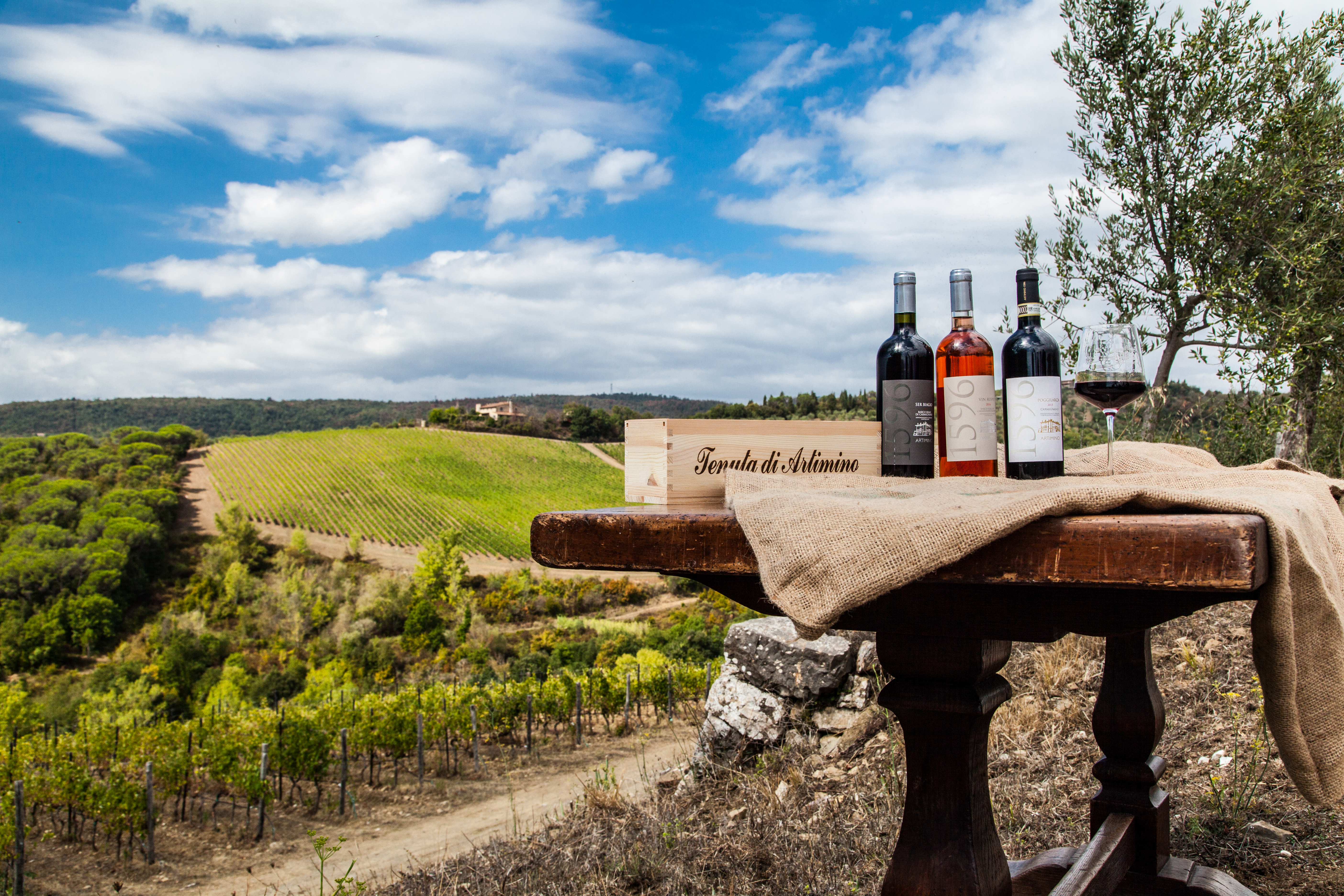 Artimino wine bottles, glass, crate on table with scenic Tuscan vineyard.