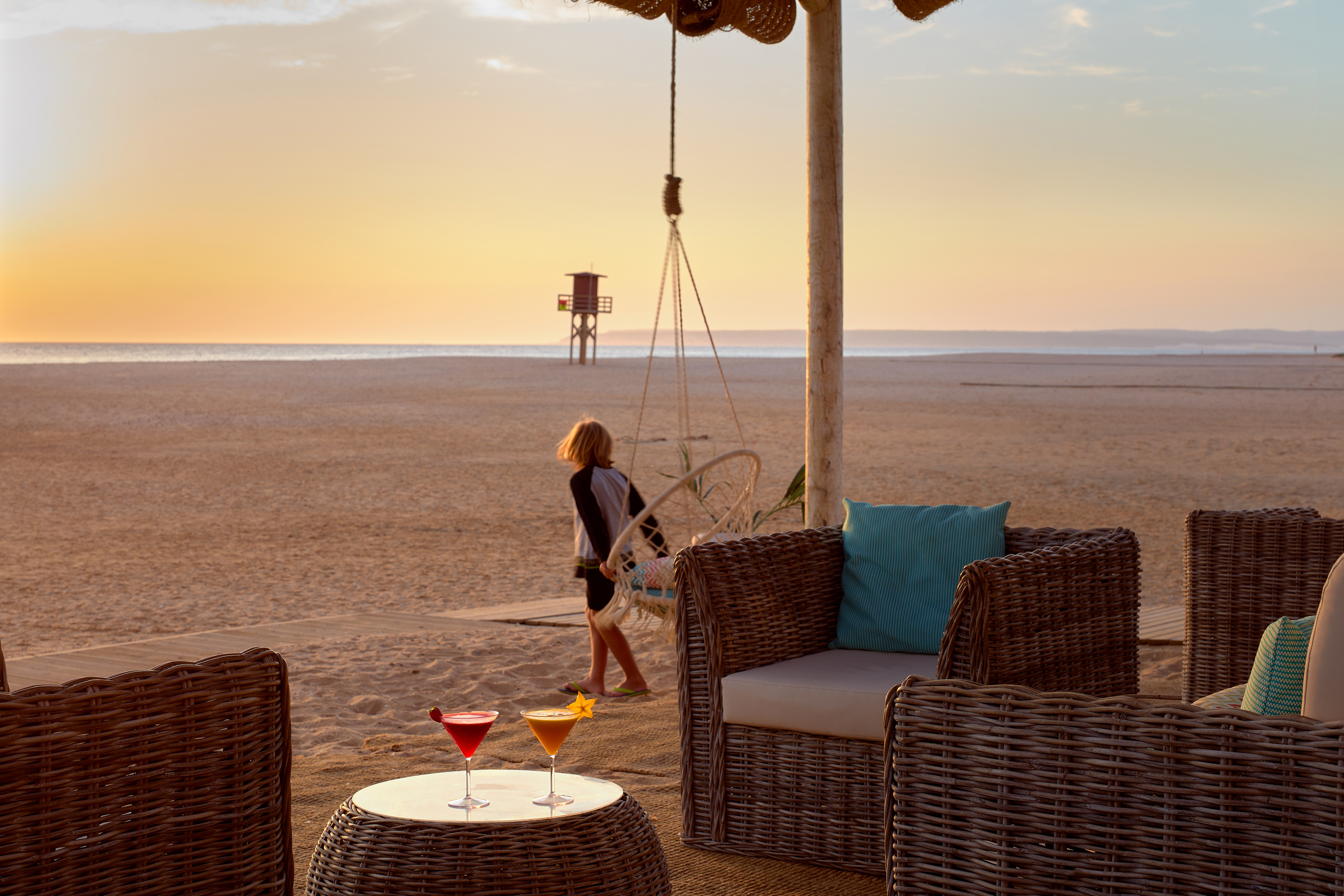 Sunset beach lounge with cocktails, wicker furniture, and child near ocean.