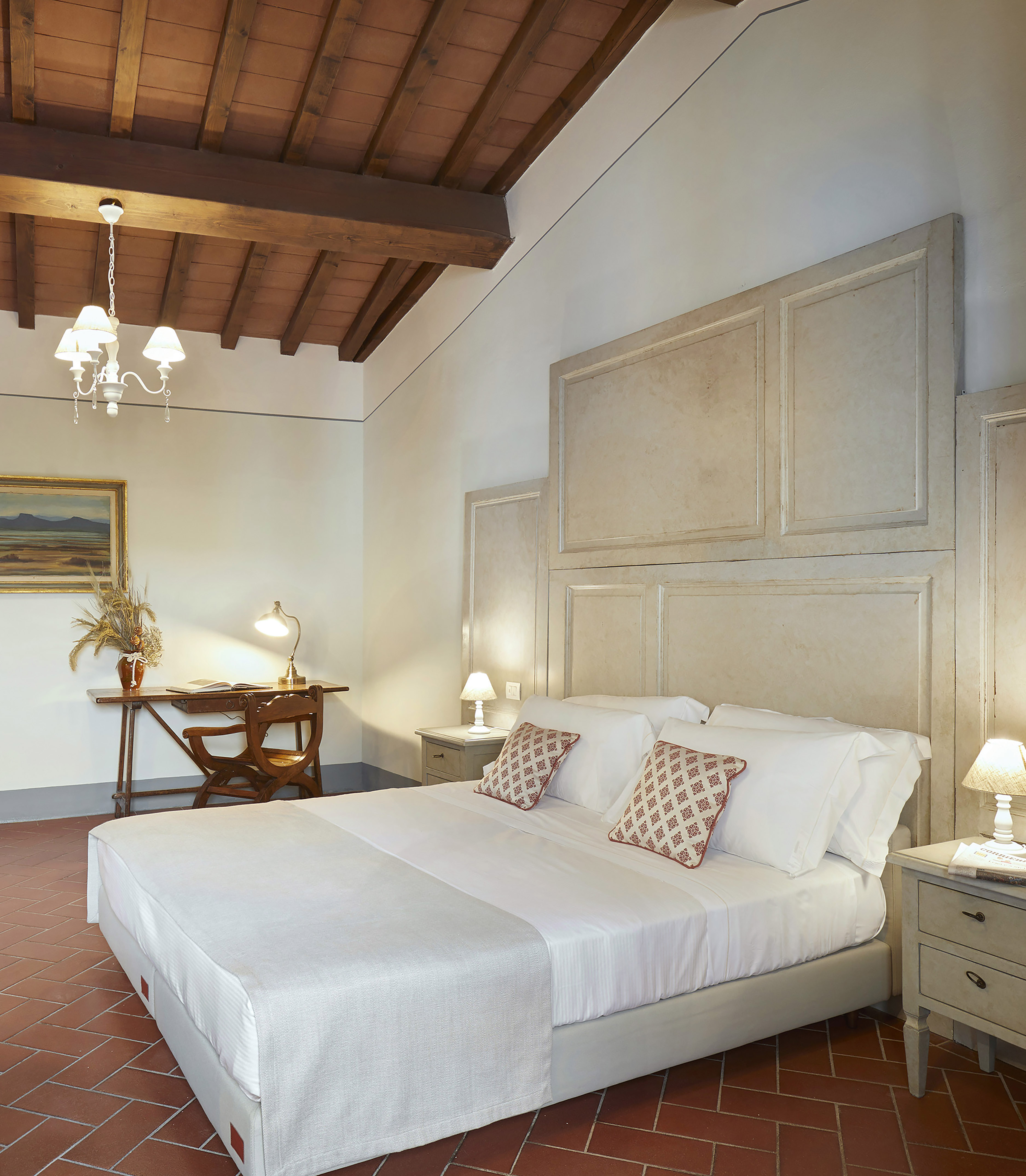Tuscan bedroom with wooden beams, terracotta floor, large bed, and reading desk.