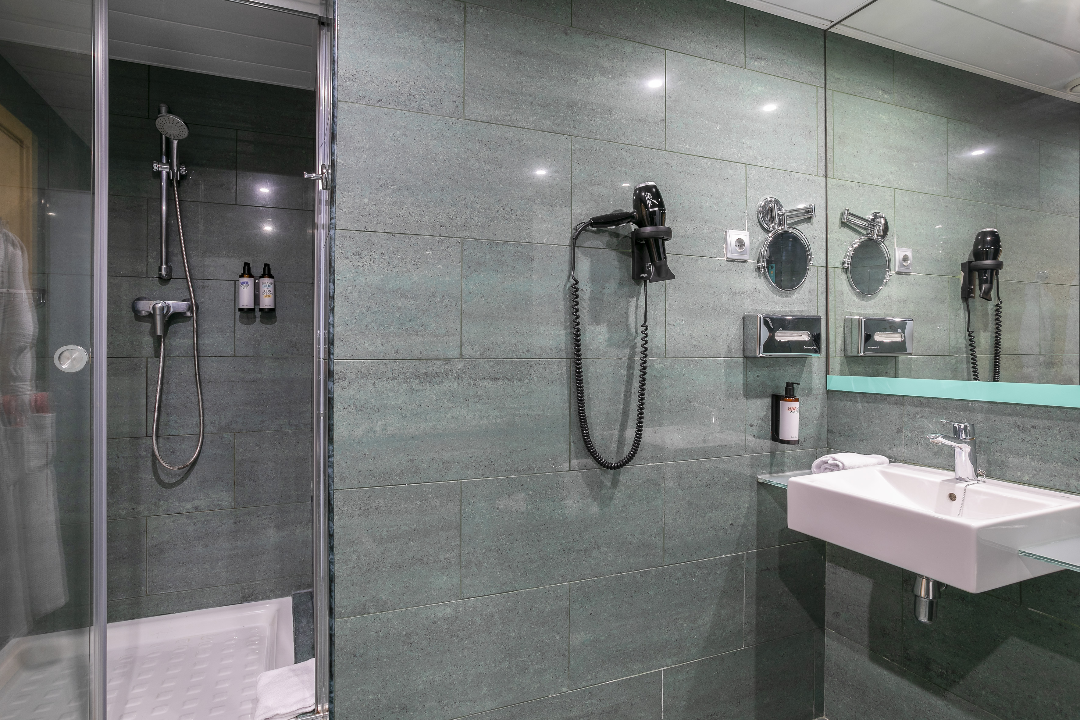 Modern hotel bathroom with shower, sink, mirror, and wall-mounted hairdryer.