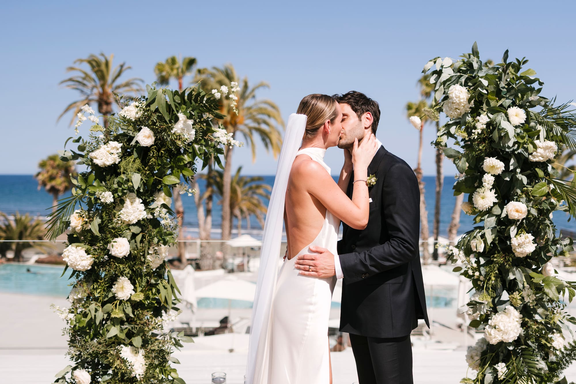 Bride and groom kissing under floral arch at tropical outdoor wedding.