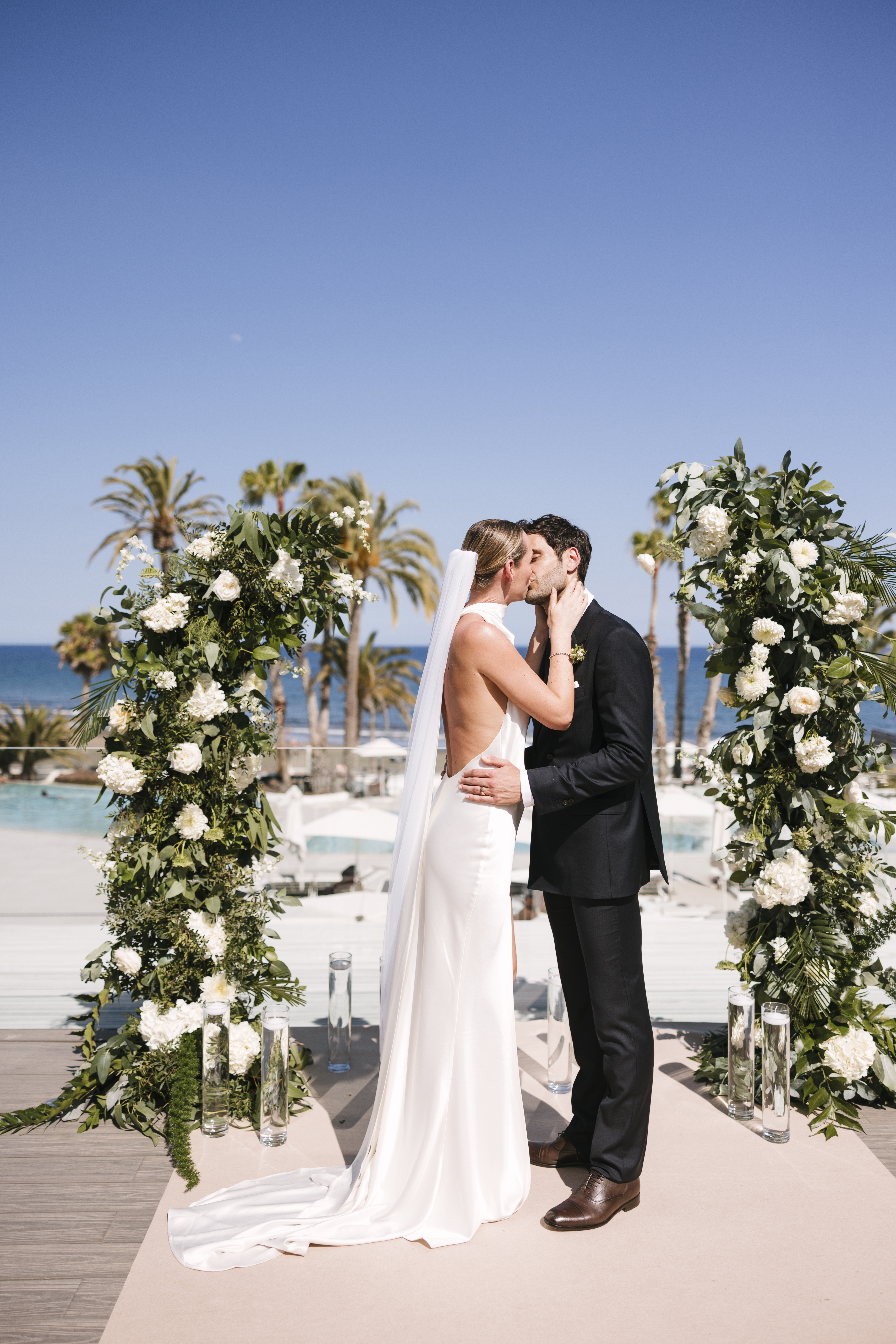 Bride and groom kissing under floral arch at tropical outdoor wedding.