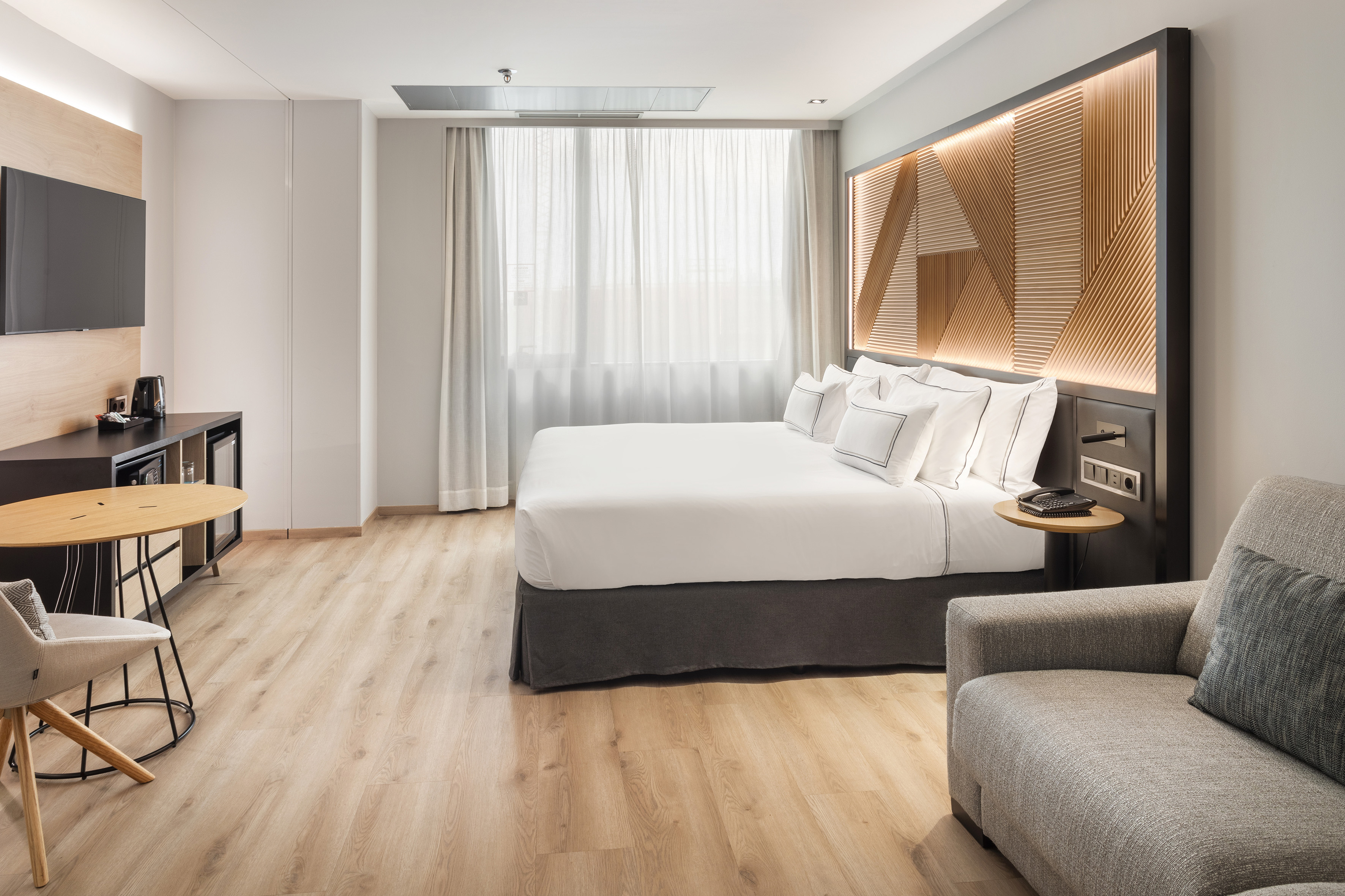 Modern hotel room with bed, lit headboard, wood floor, TV, and seating.
