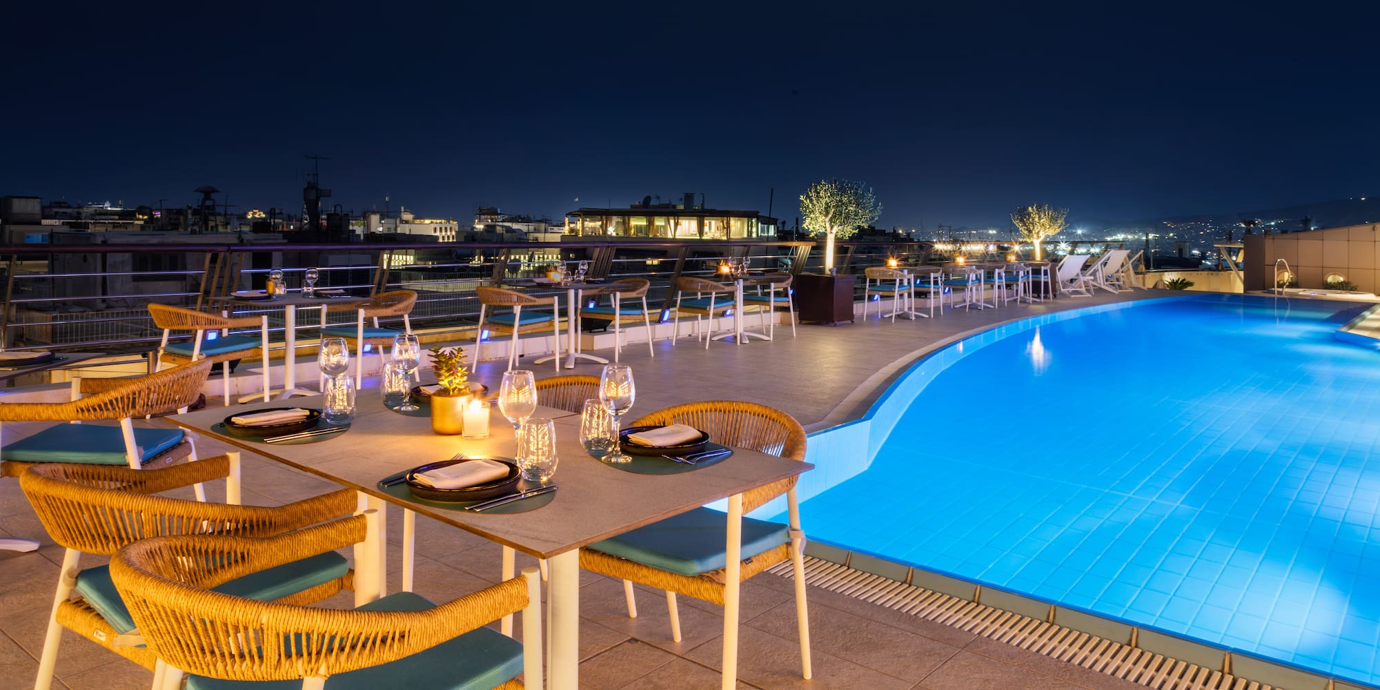 Rooftop dining, candlelit tables, sparkling blue pool, and night city view.