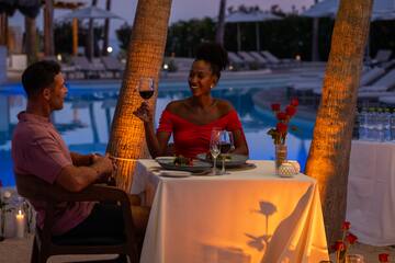 a woman holding a glass of wine at a table with a man sitting by a pool