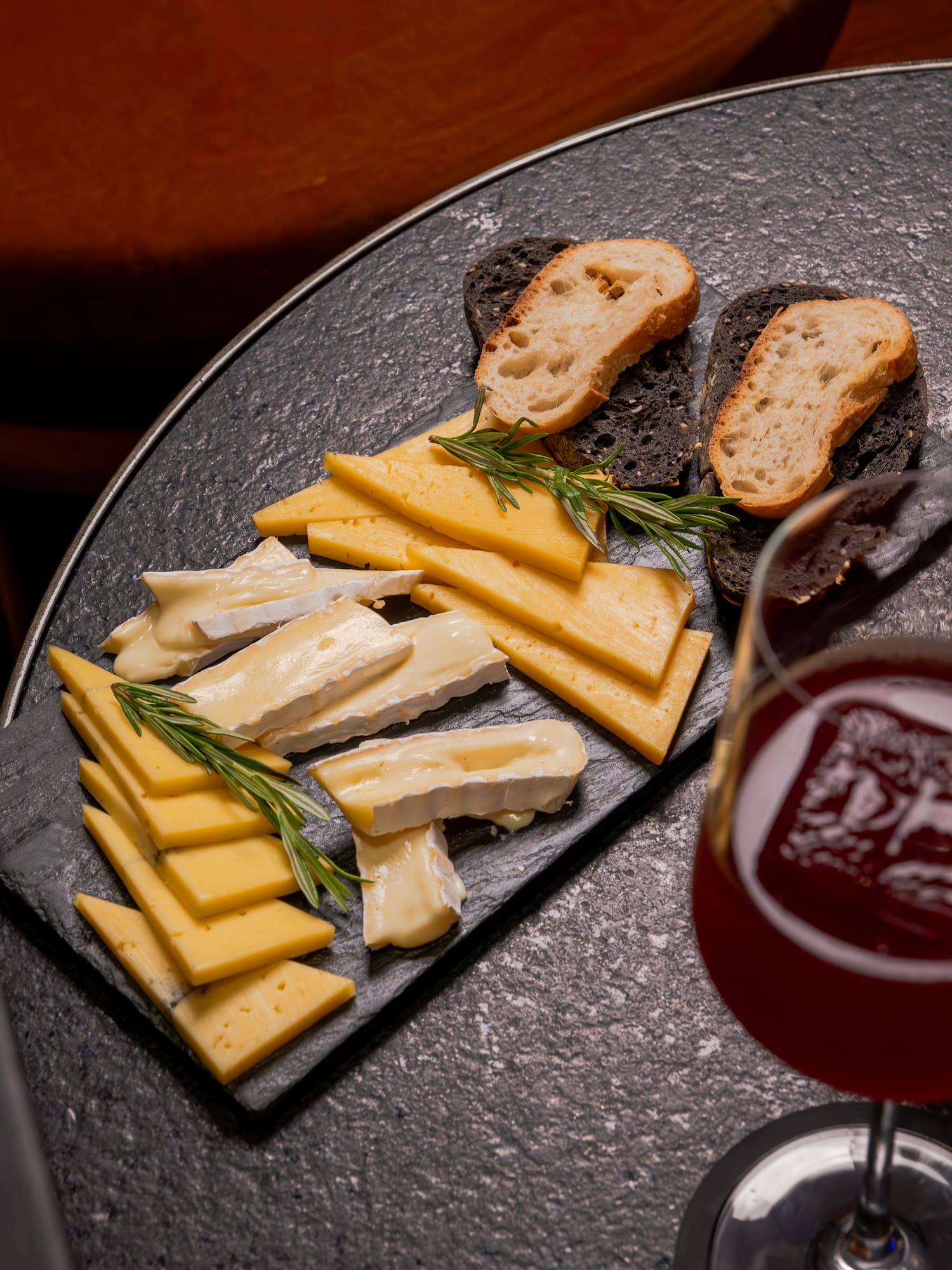 Tabla de quesos, pan y romero con vino tinto en mesa oscura.