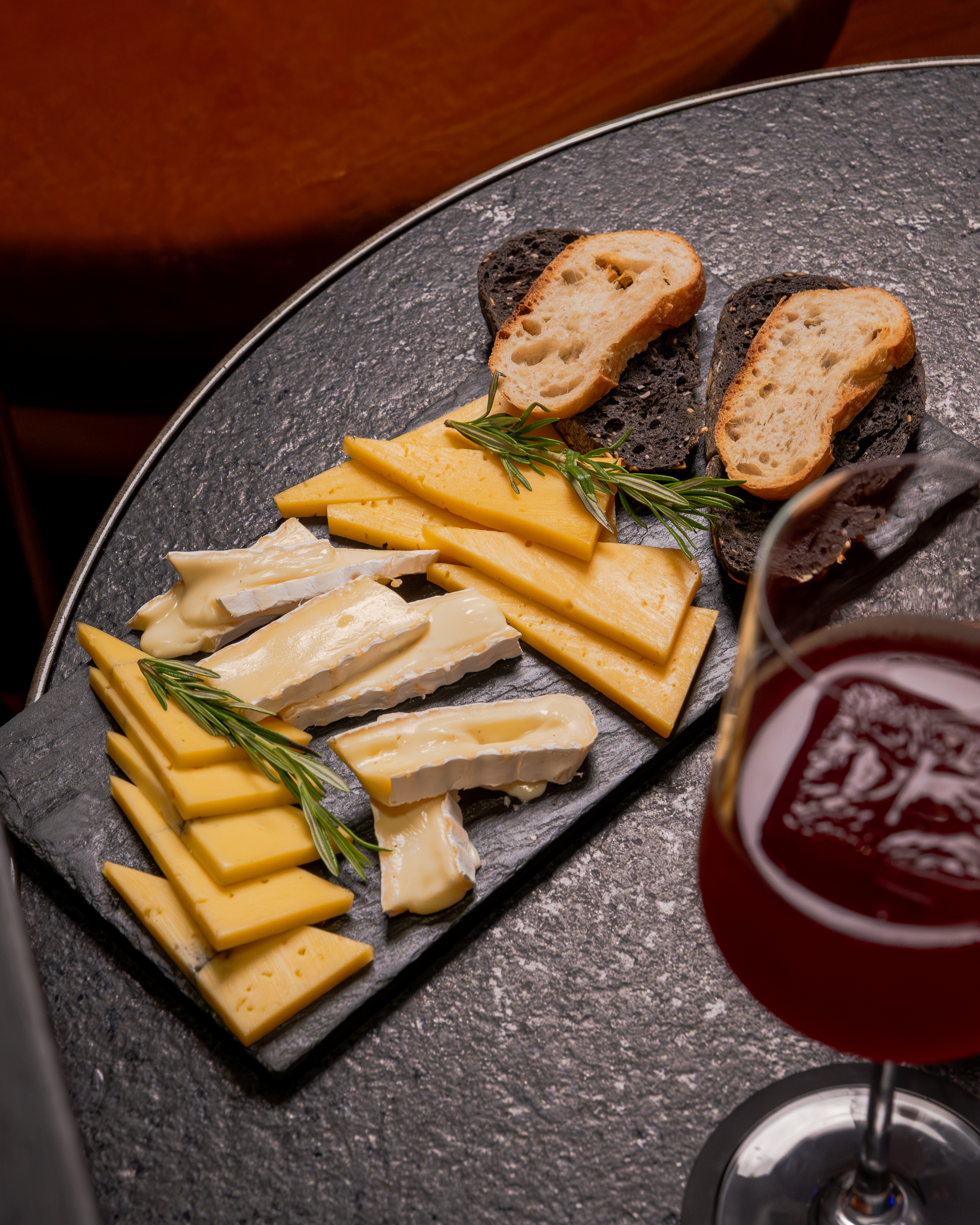 Tabla de quesos, pan y romero con vino tinto en mesa oscura.