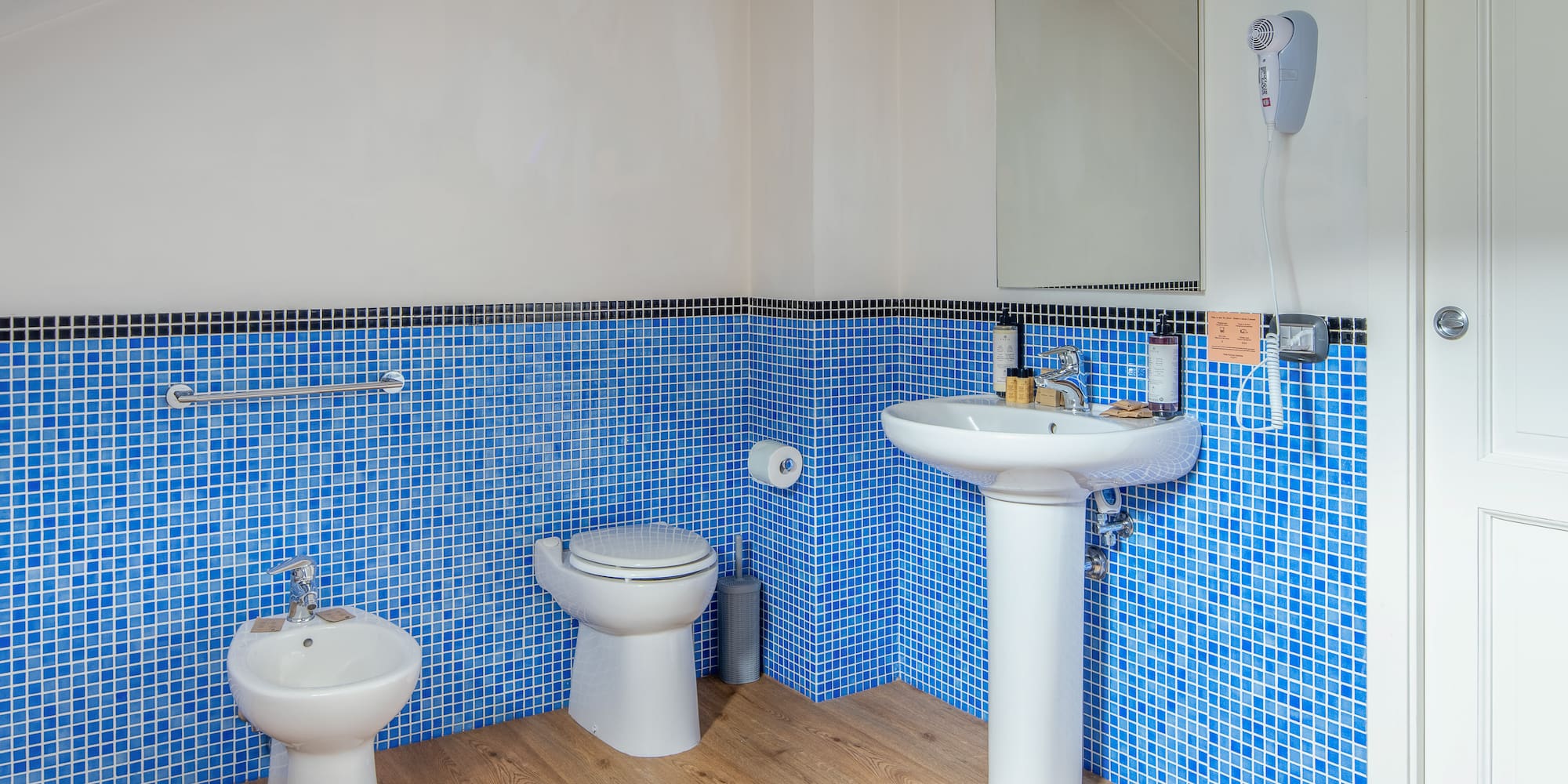 Blue mosaic tile bathroom with toilet, bidet, sink, and wooden floor.