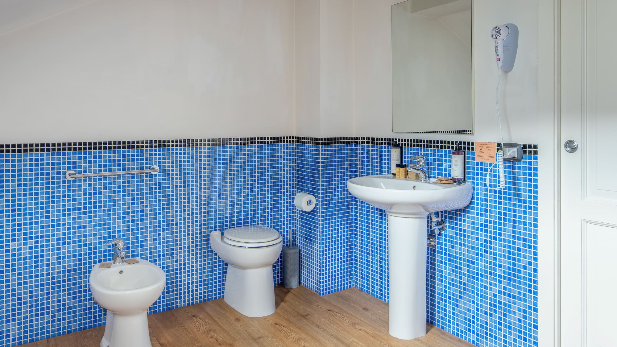 Blue mosaic tile bathroom with toilet, bidet, sink, and wooden floor.