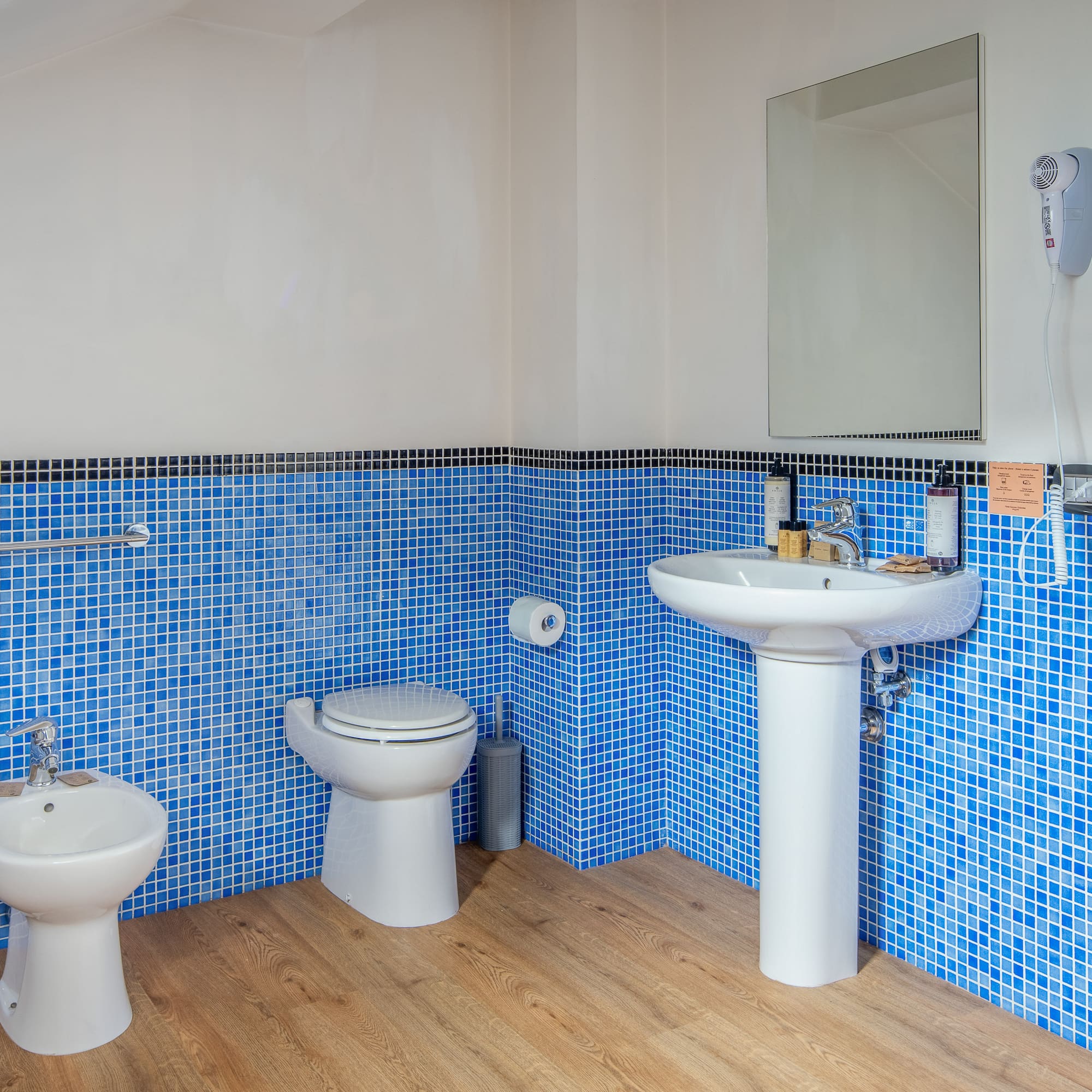 Blue mosaic tile bathroom with toilet, bidet, sink, and wooden floor.