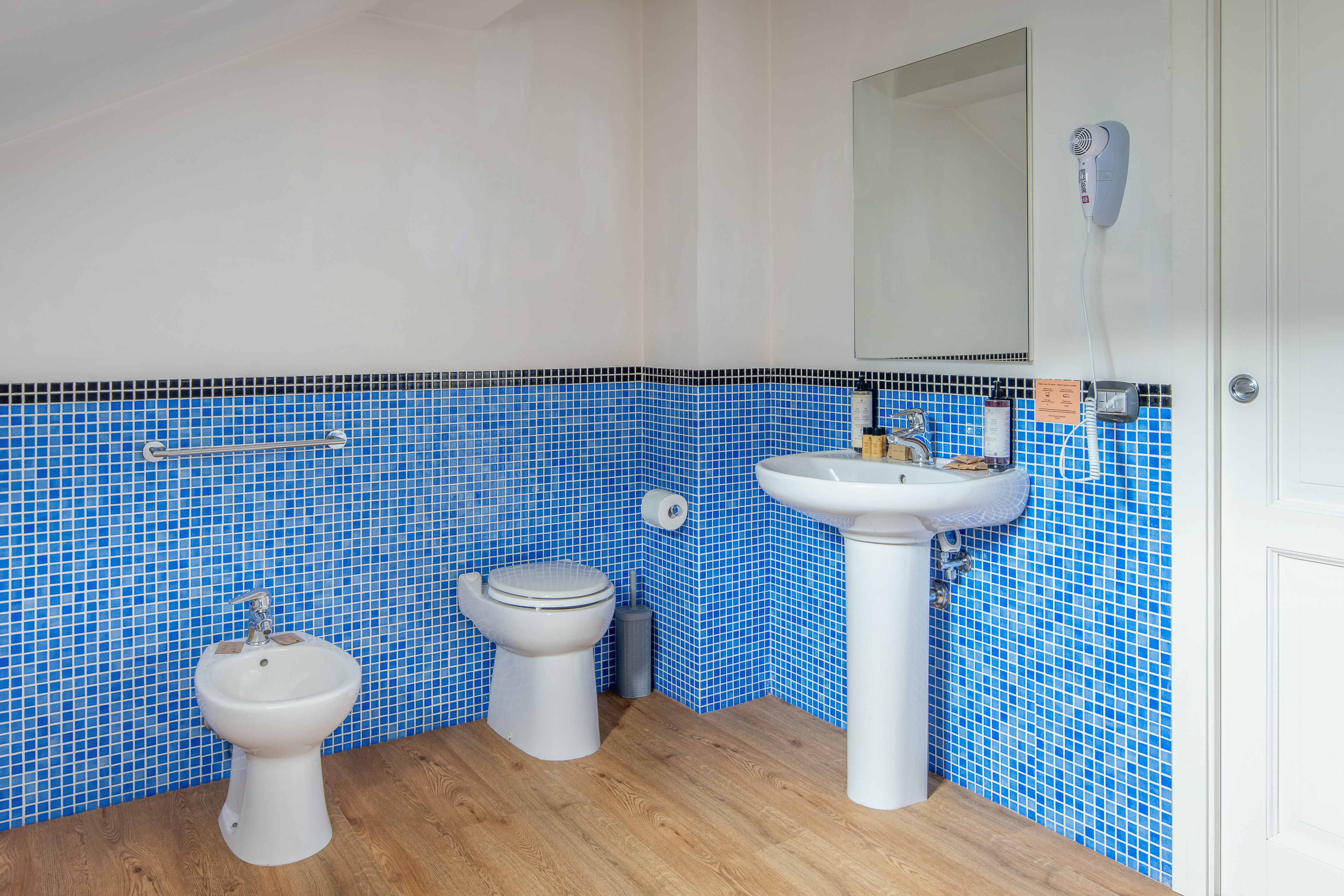 Blue mosaic tile bathroom with toilet, bidet, sink, and wooden floor.