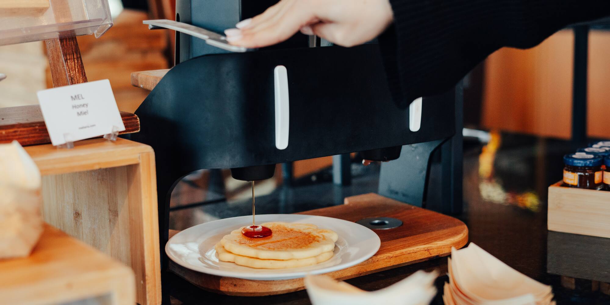 Person's hand dispensing honey onto fresh pancakes at a breakfast buffet.