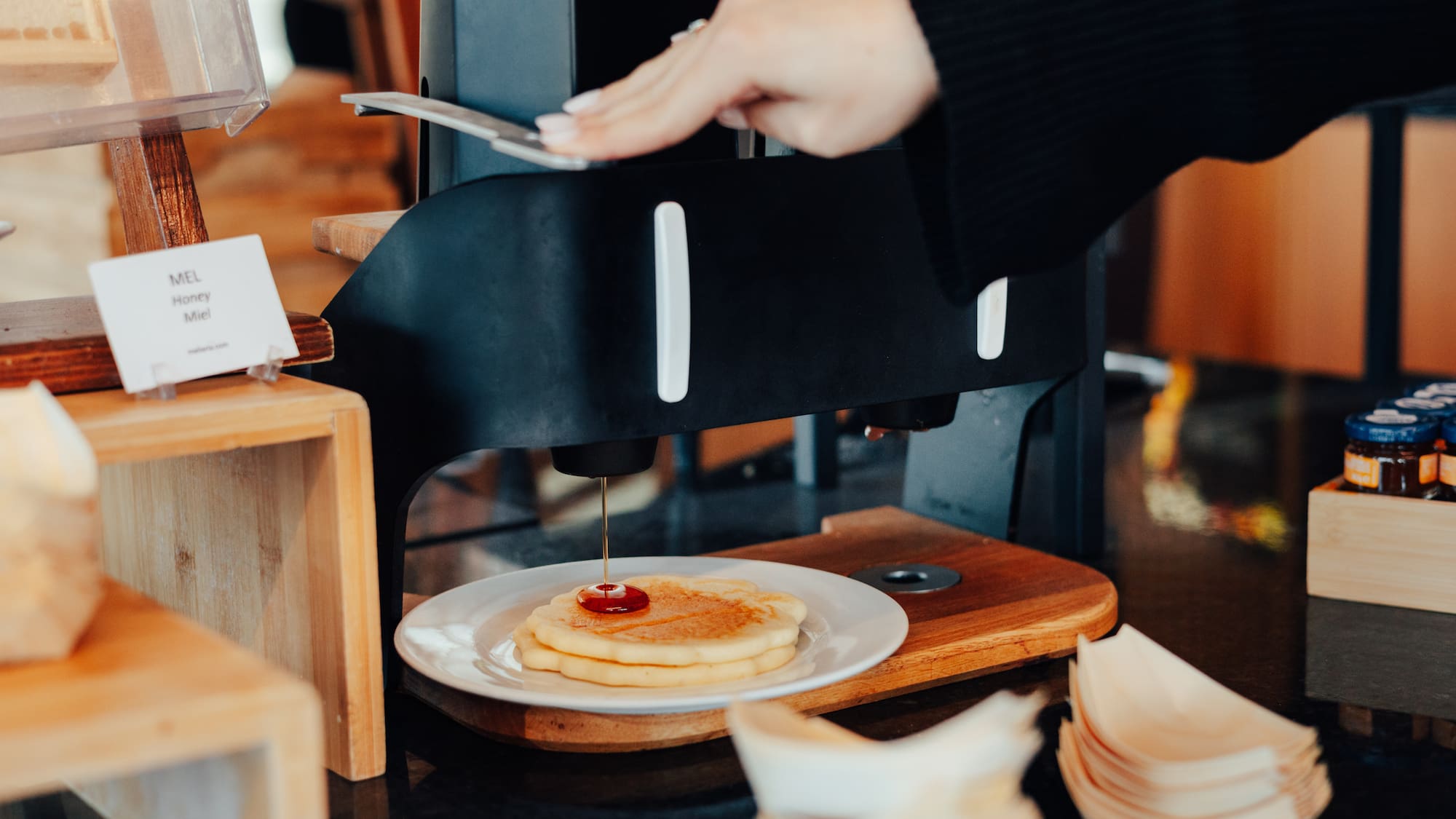 Person's hand dispensing honey onto fresh pancakes at a breakfast buffet.