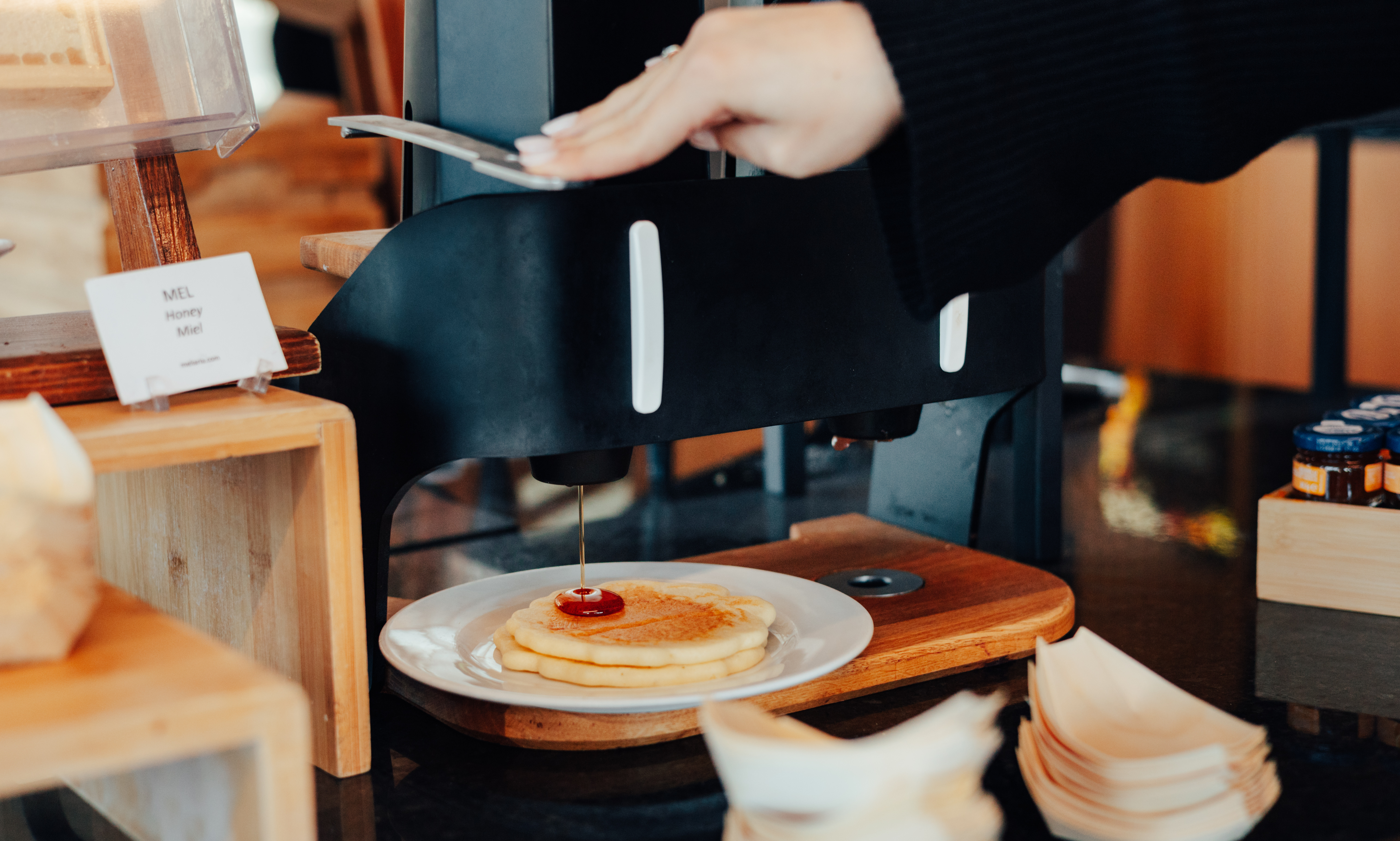 Person's hand dispensing honey onto fresh pancakes at a breakfast buffet.