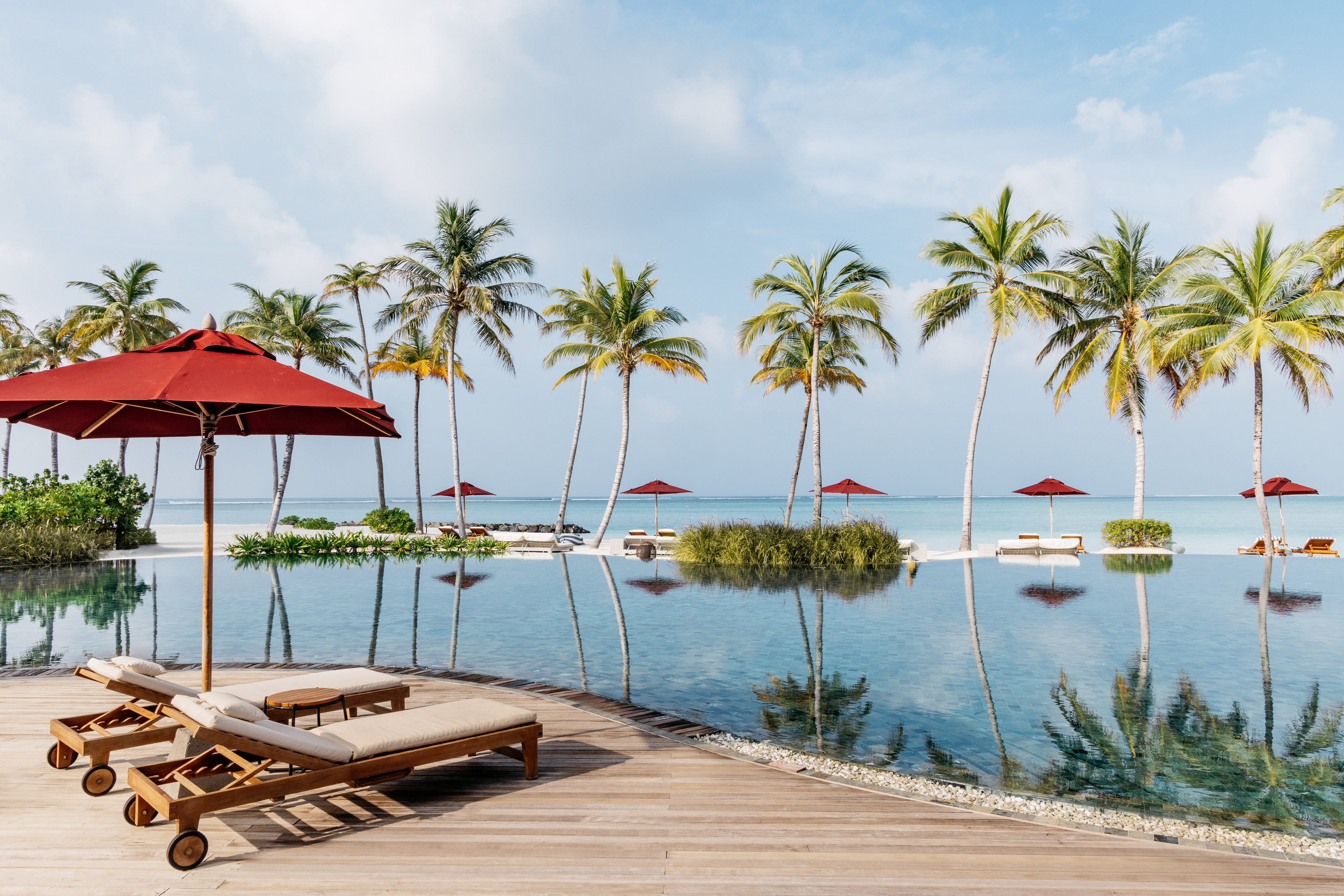 Luxury resort infinity pool with palm trees, red umbrellas, and ocean view.