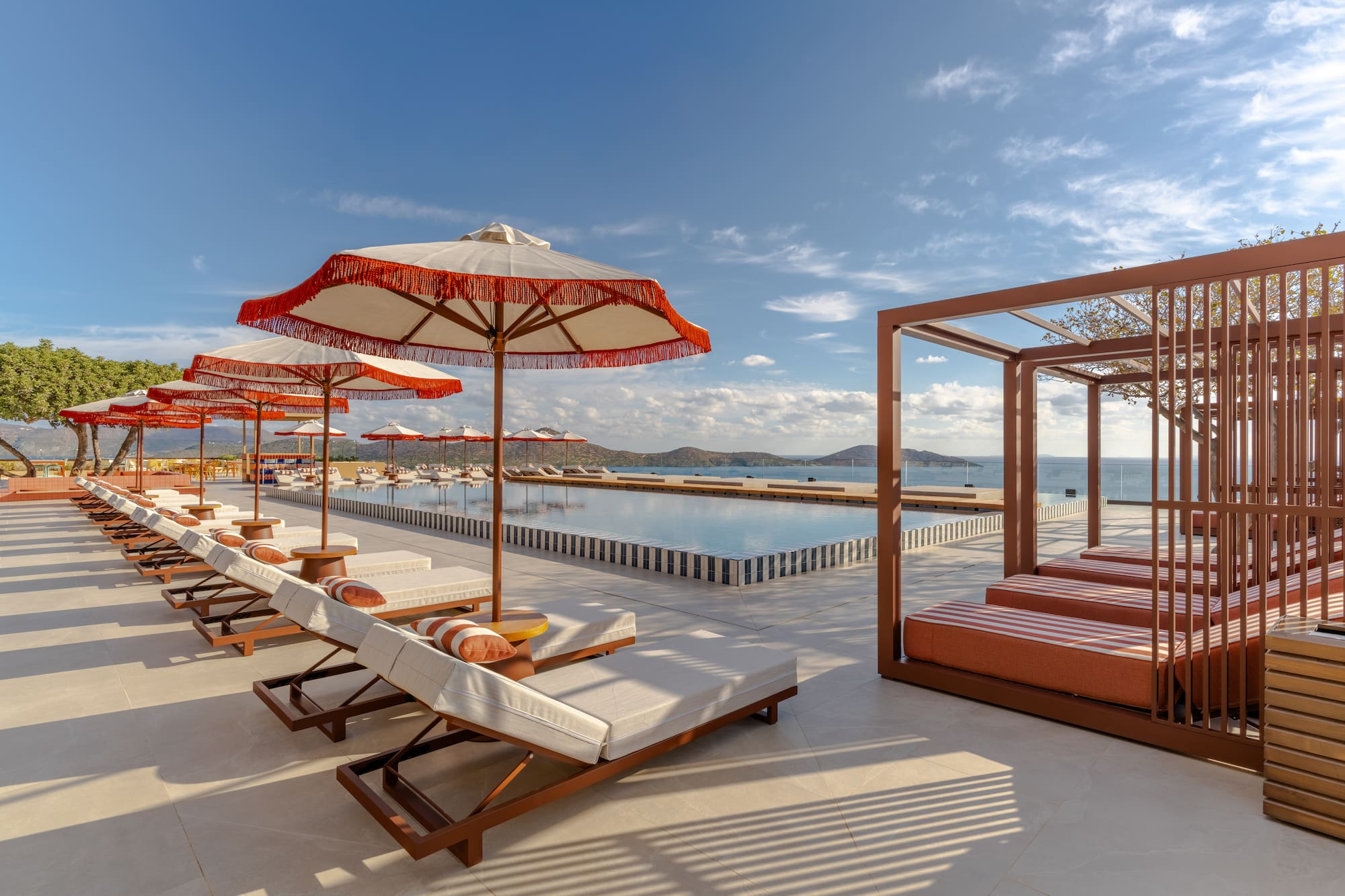 Luxury resort infinity pool, ocean view, sunbeds, and red umbrellas.