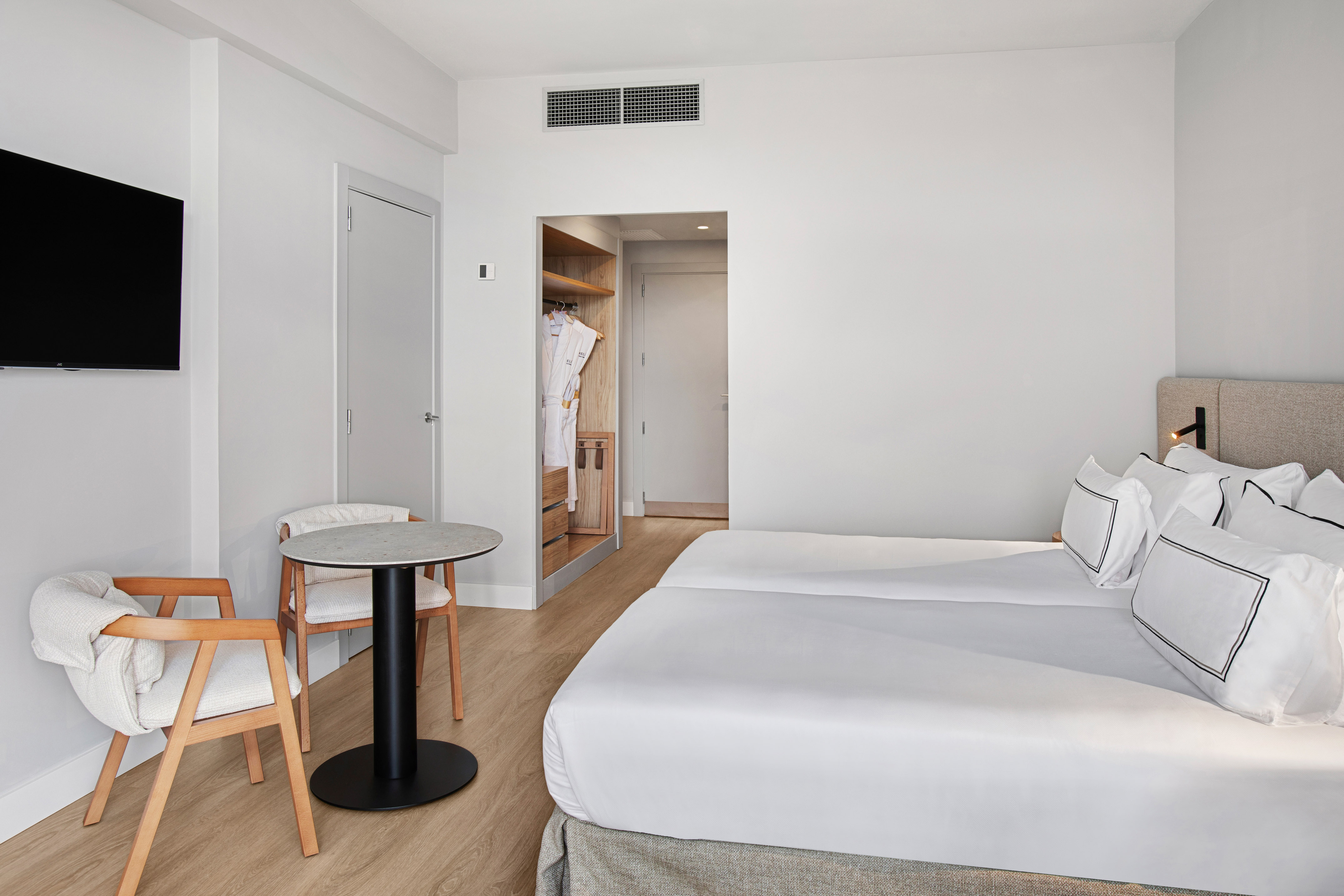 Modern hotel room with twin beds, white decor, wood floor, TV, and open closet.