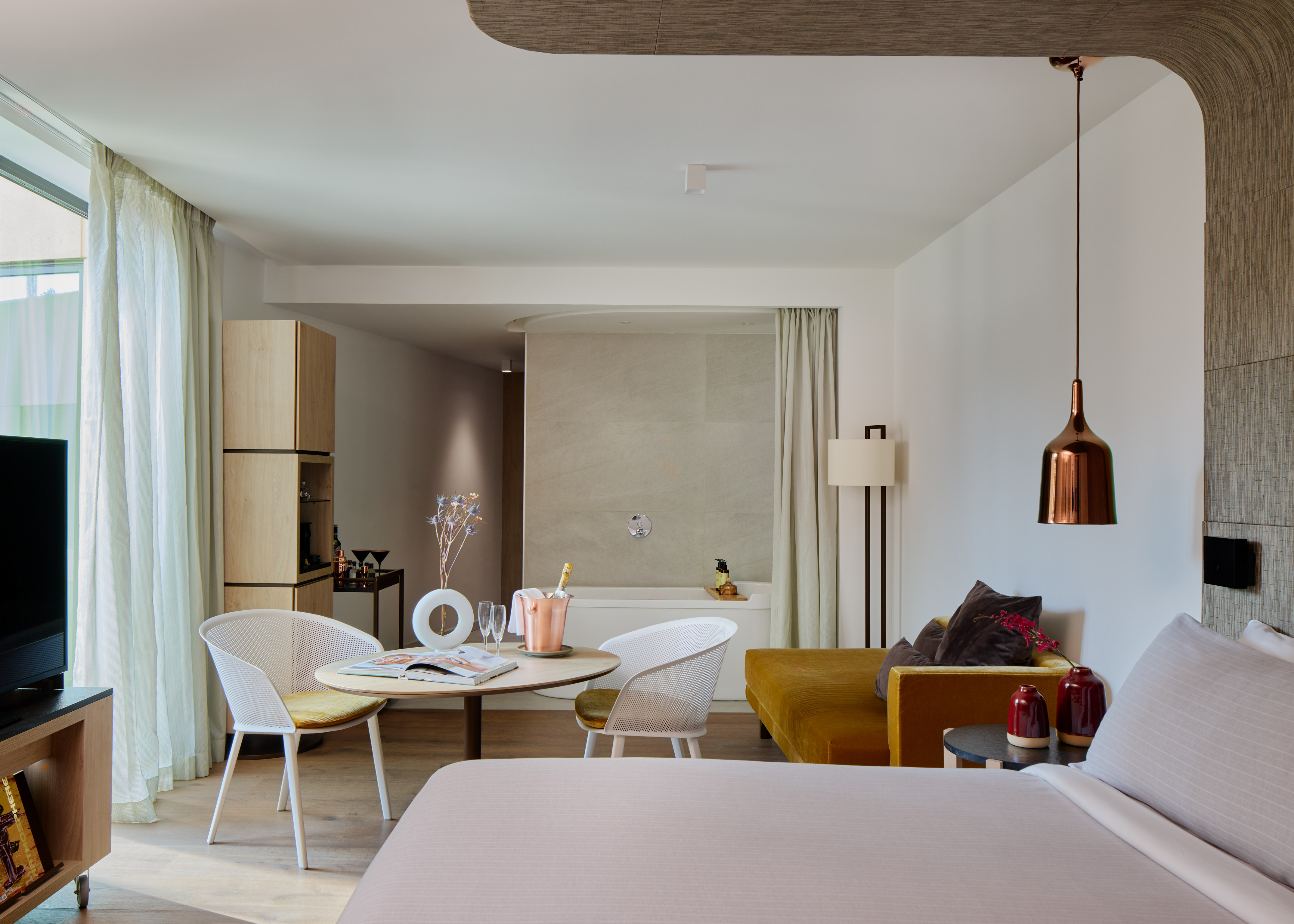 Modern luxury hotel suite with bed, dining table, sofa, and open bathtub.