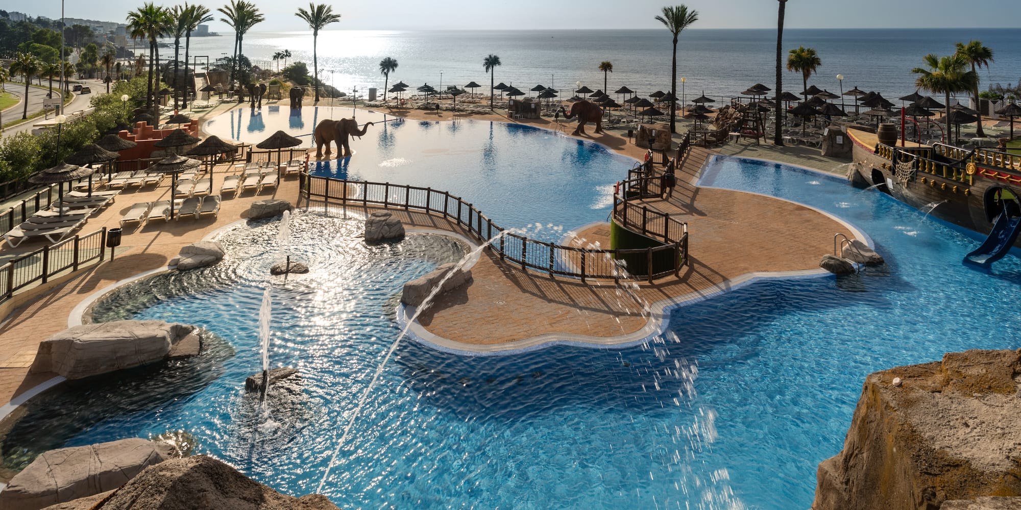Resort pool with elephant statues, pirate ship slide, palm trees, and ocean view.