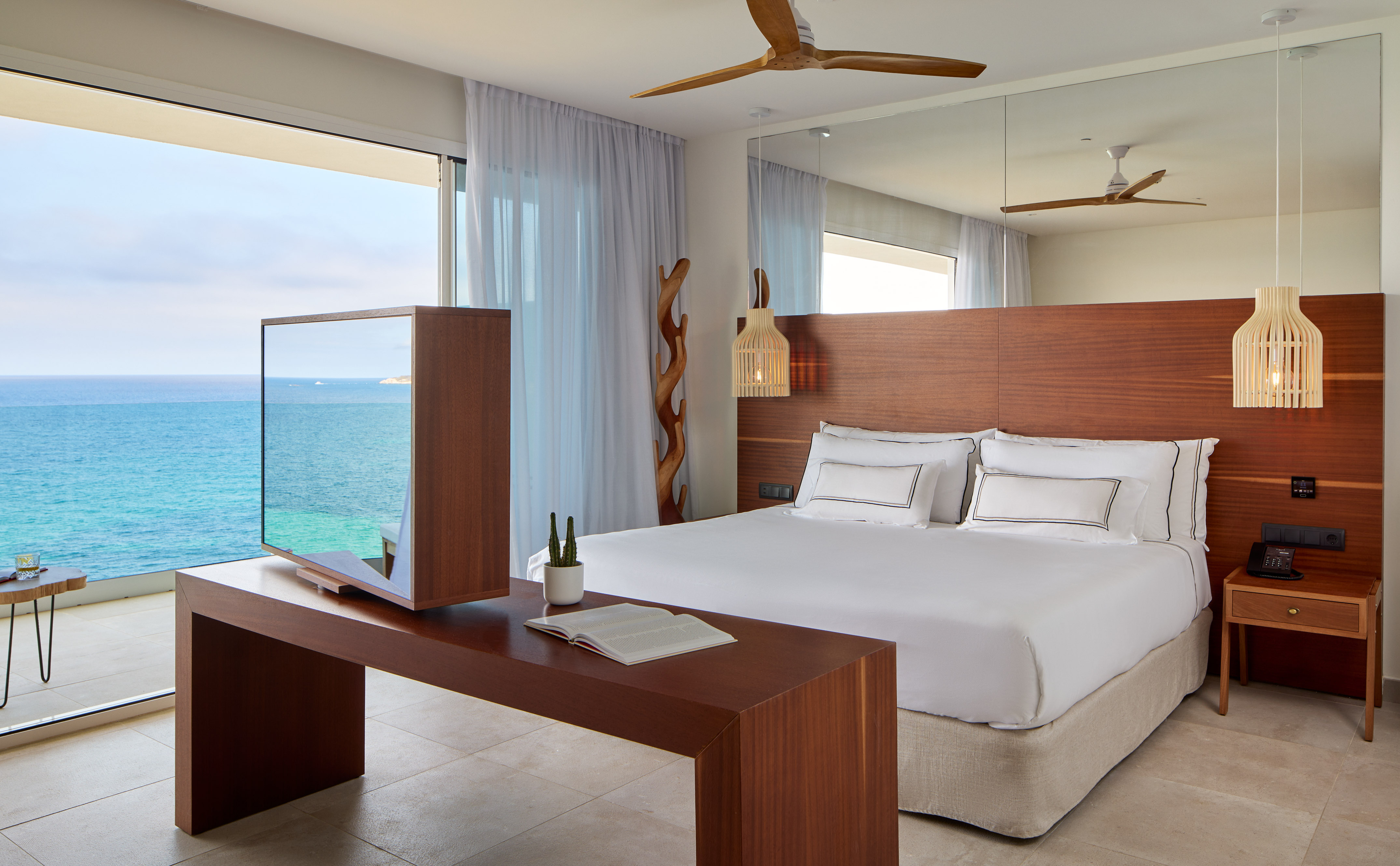 Modern hotel bedroom with panoramic ocean view, wooden headboard, and rotating TV.