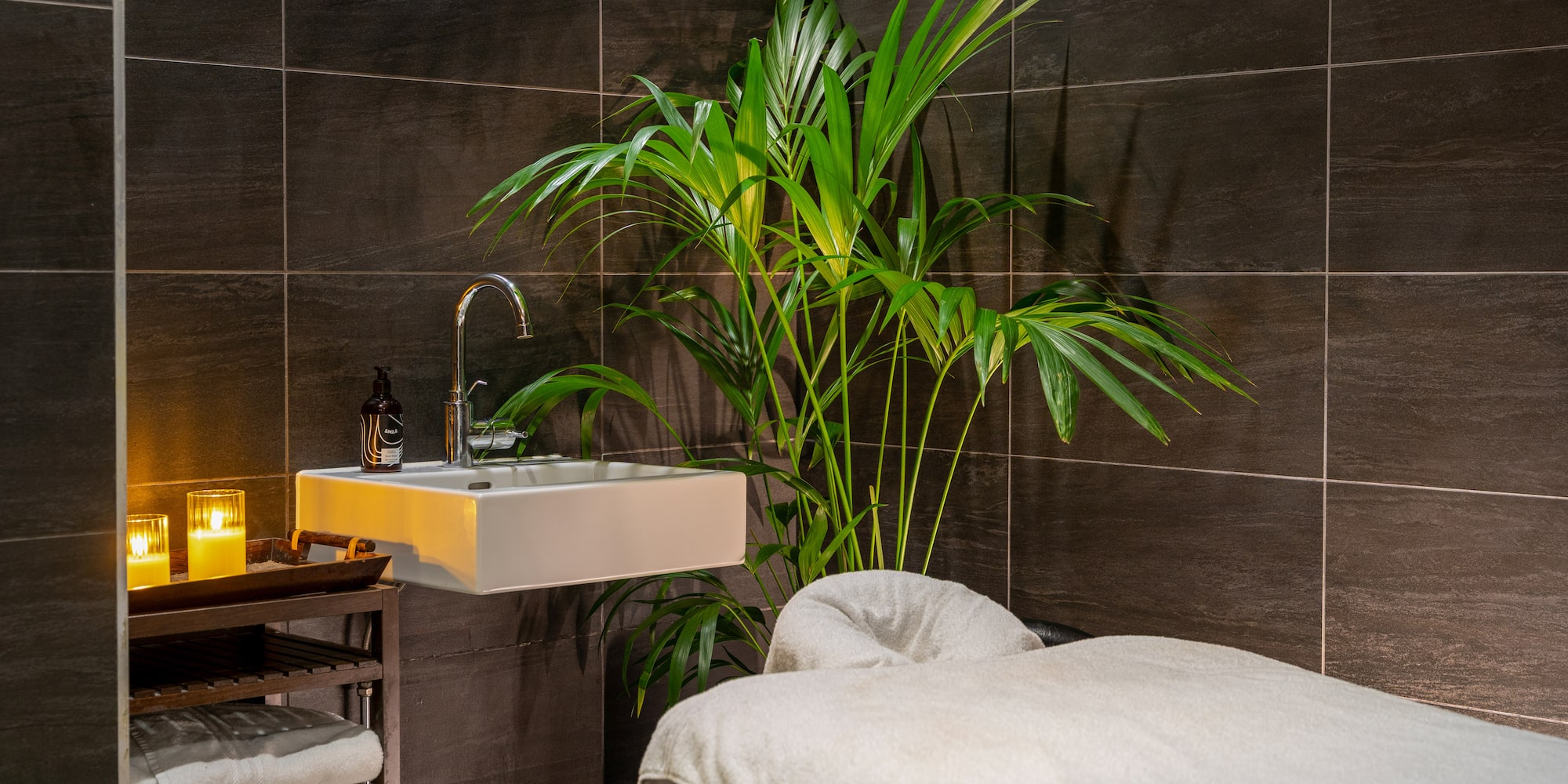 Luxurious spa treatment room with massage table, sink, palm plant, and lit candles.