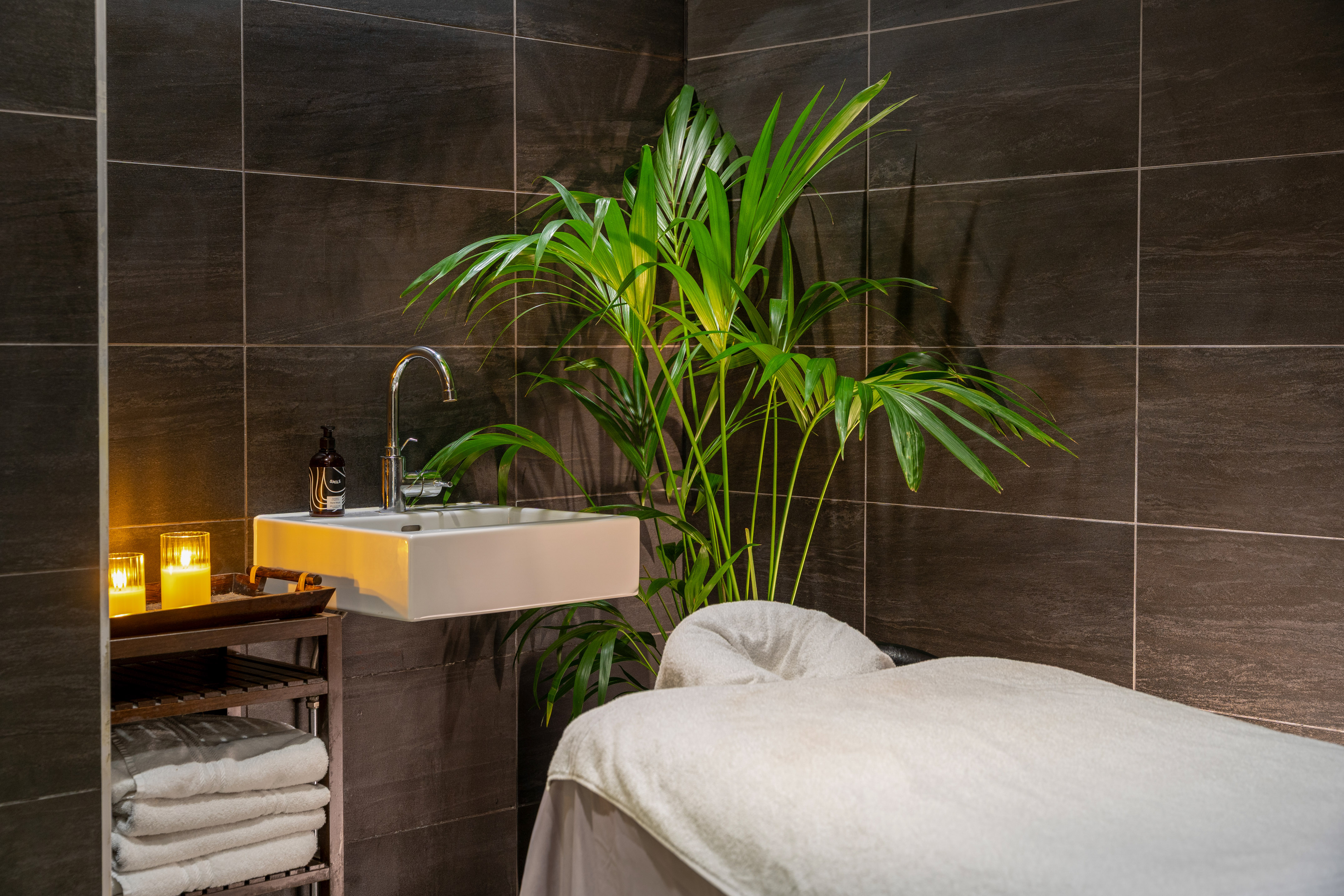Luxurious spa treatment room with massage table, sink, palm plant, and lit candles.