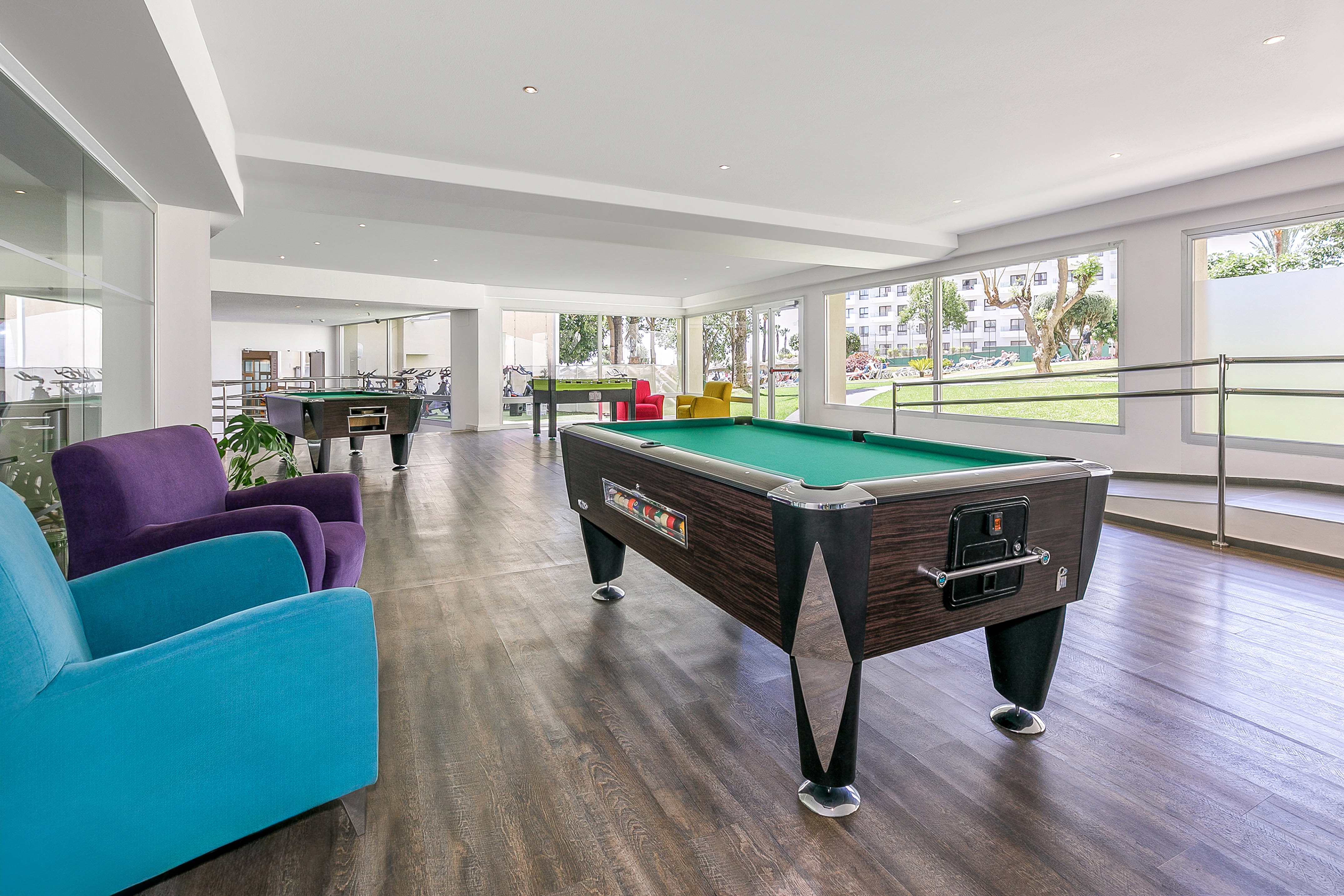 Hotel game room with pool tables, foosball, colorful seating, and large windows.