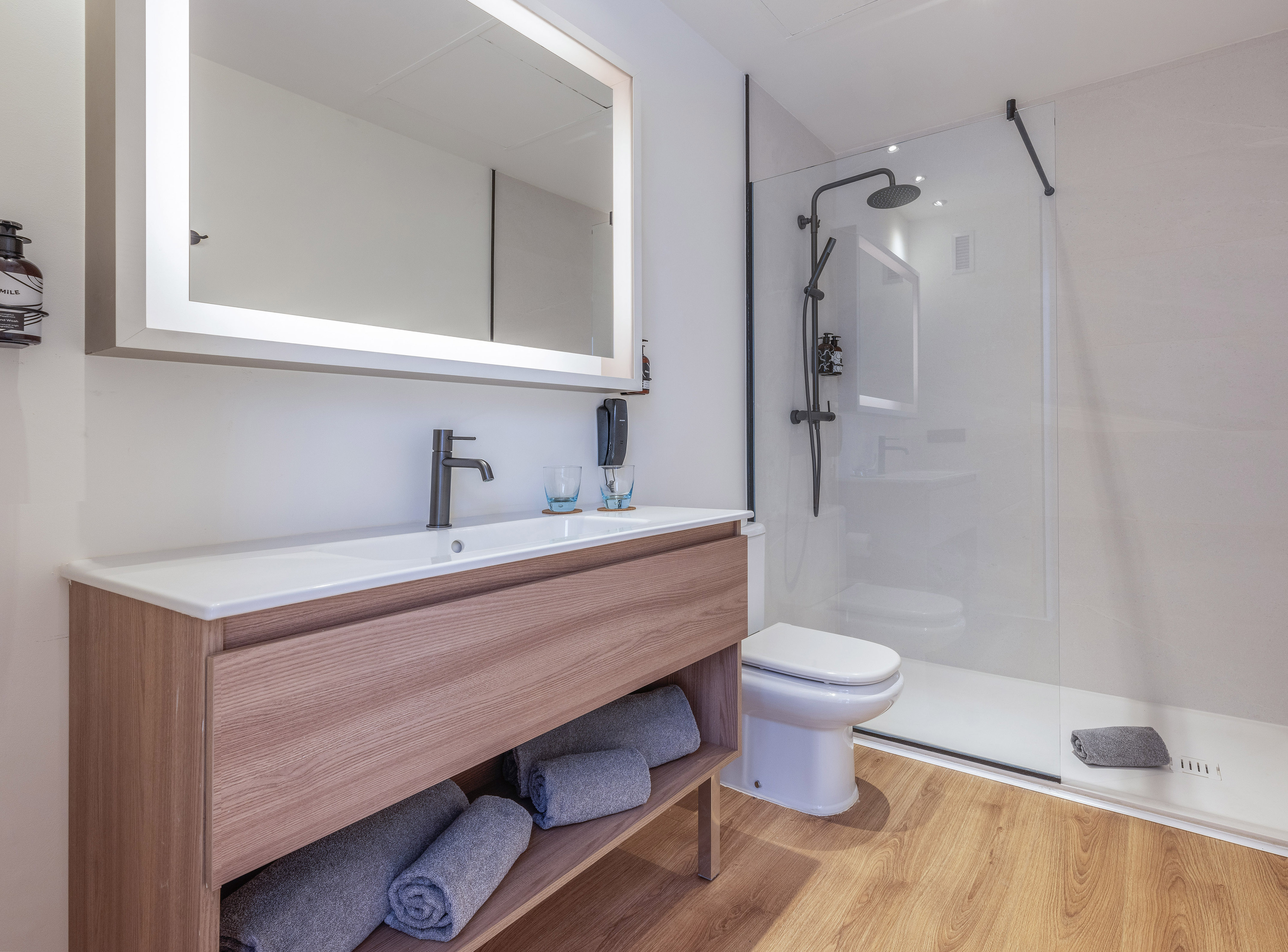 Modern hotel bathroom with wood vanity, illuminated mirror, and walk-in shower.