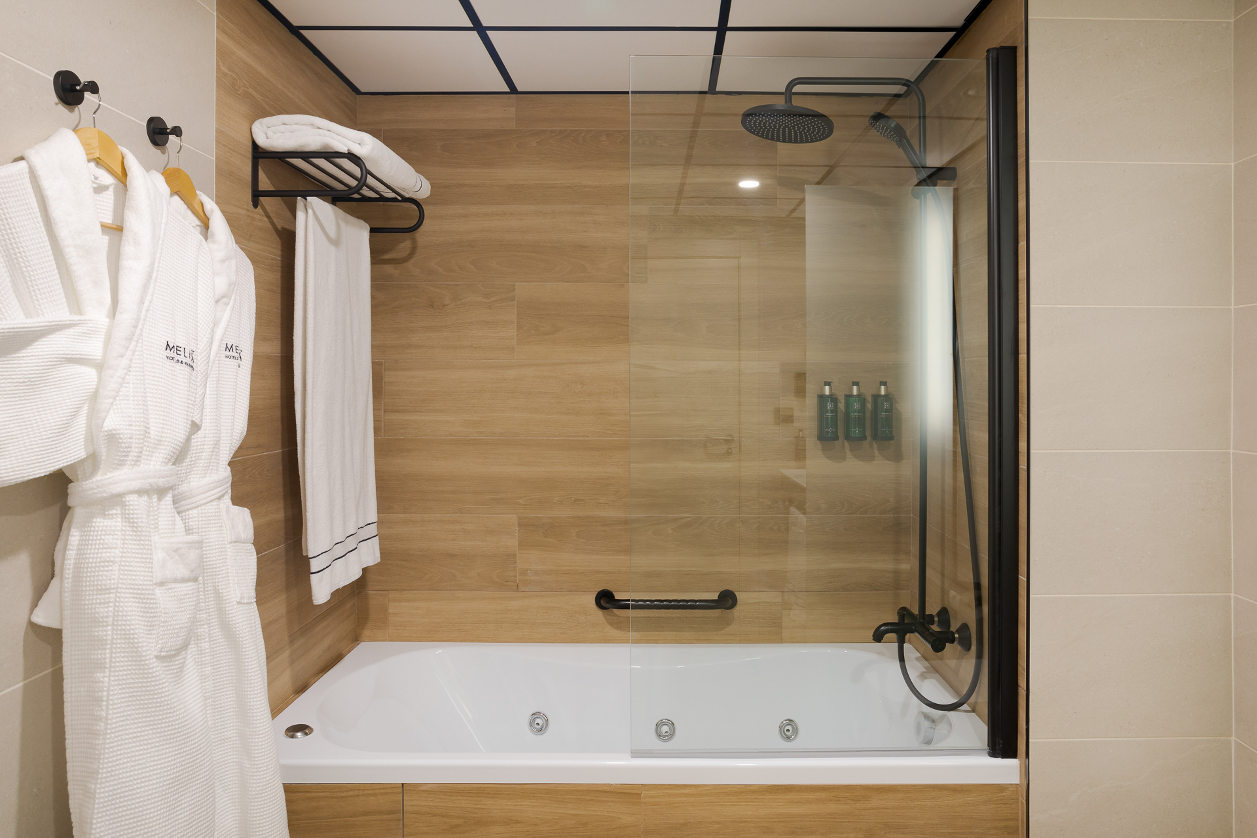Modern hotel bathroom with bathtub, glass shower, wood paneling, and bathrobes.