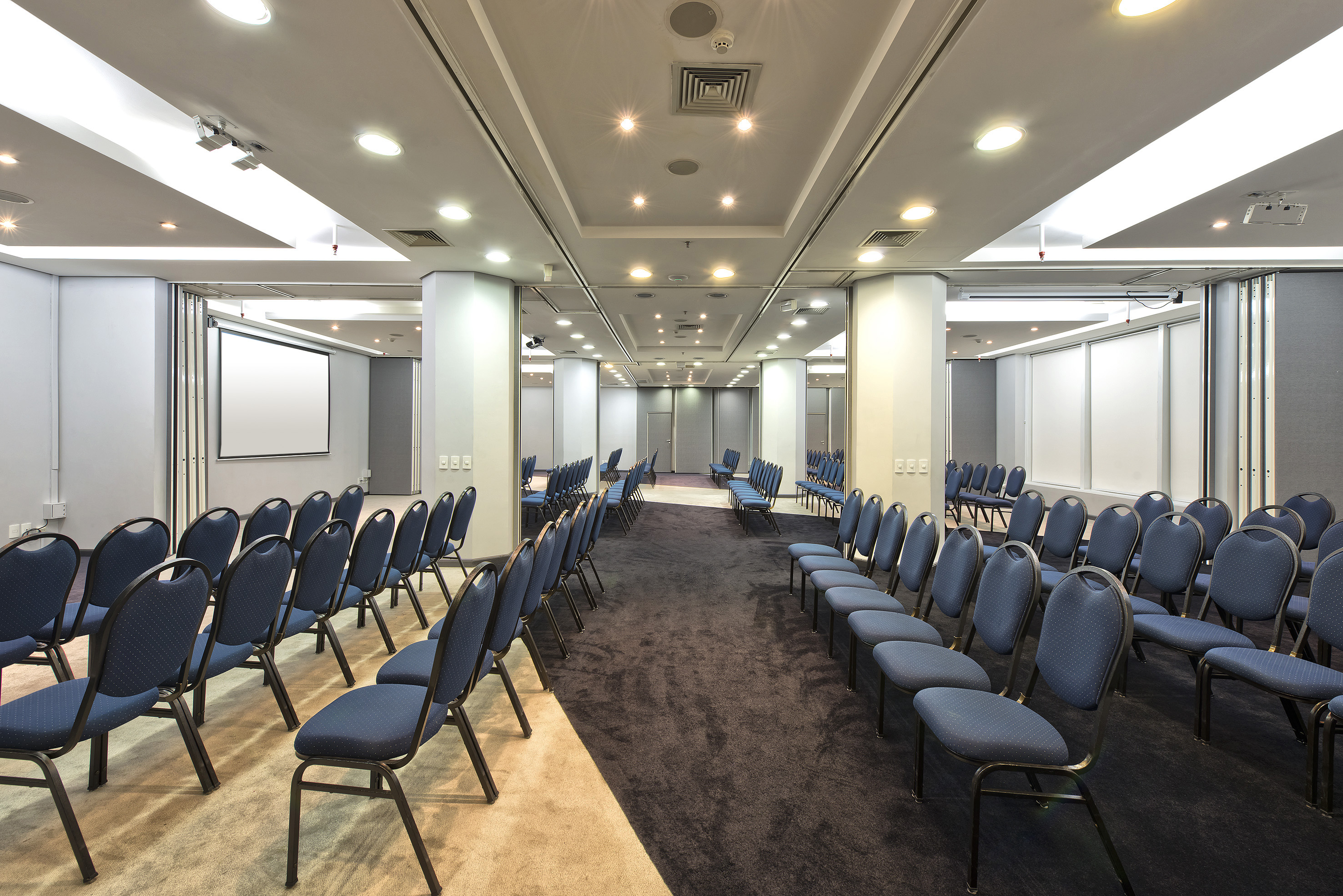 Empty conference room with rows of blue chairs, a projection screen, and bright lighting.