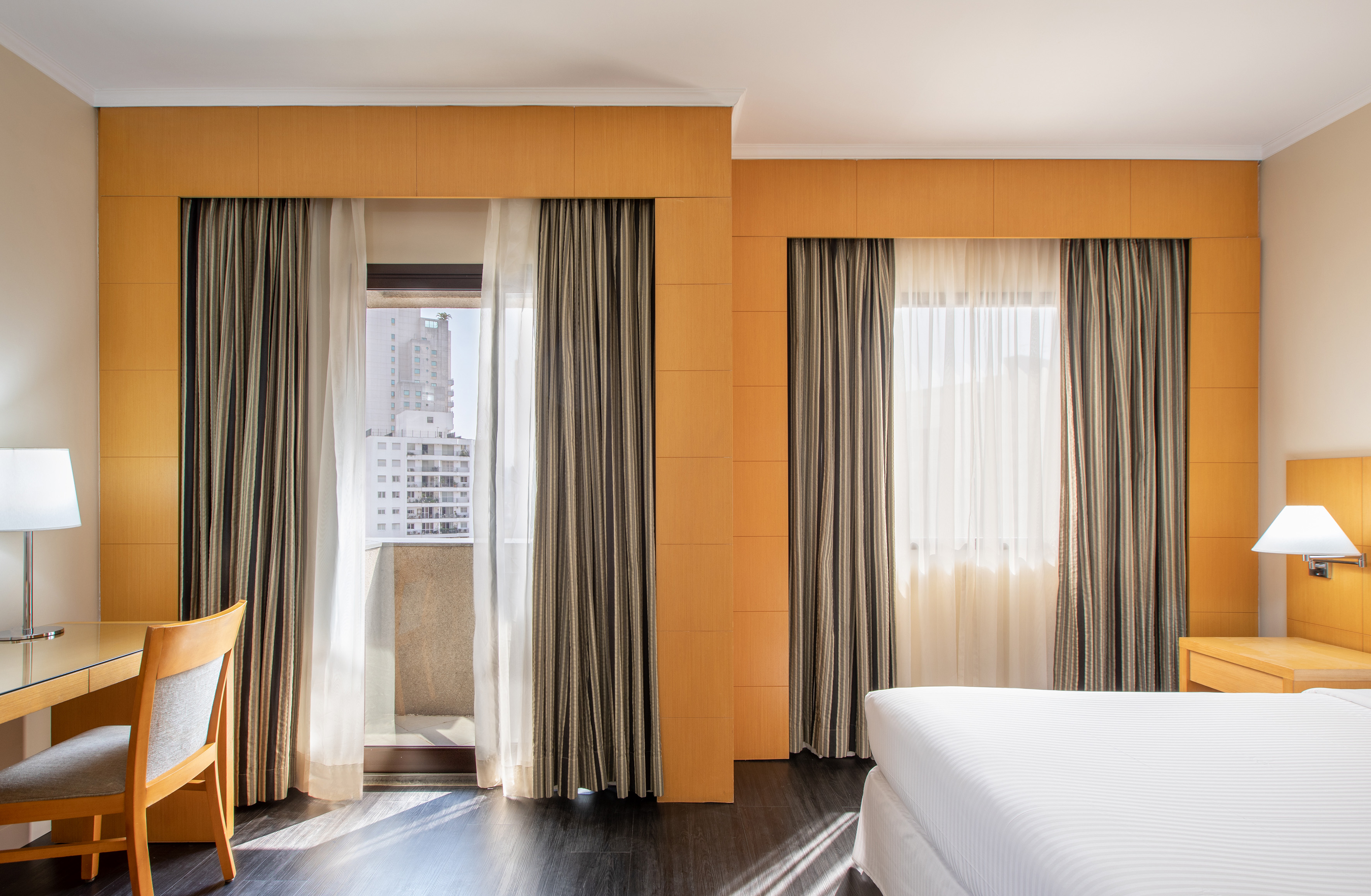 Modern hotel room with desk, bed, large windows, city view, and balcony.