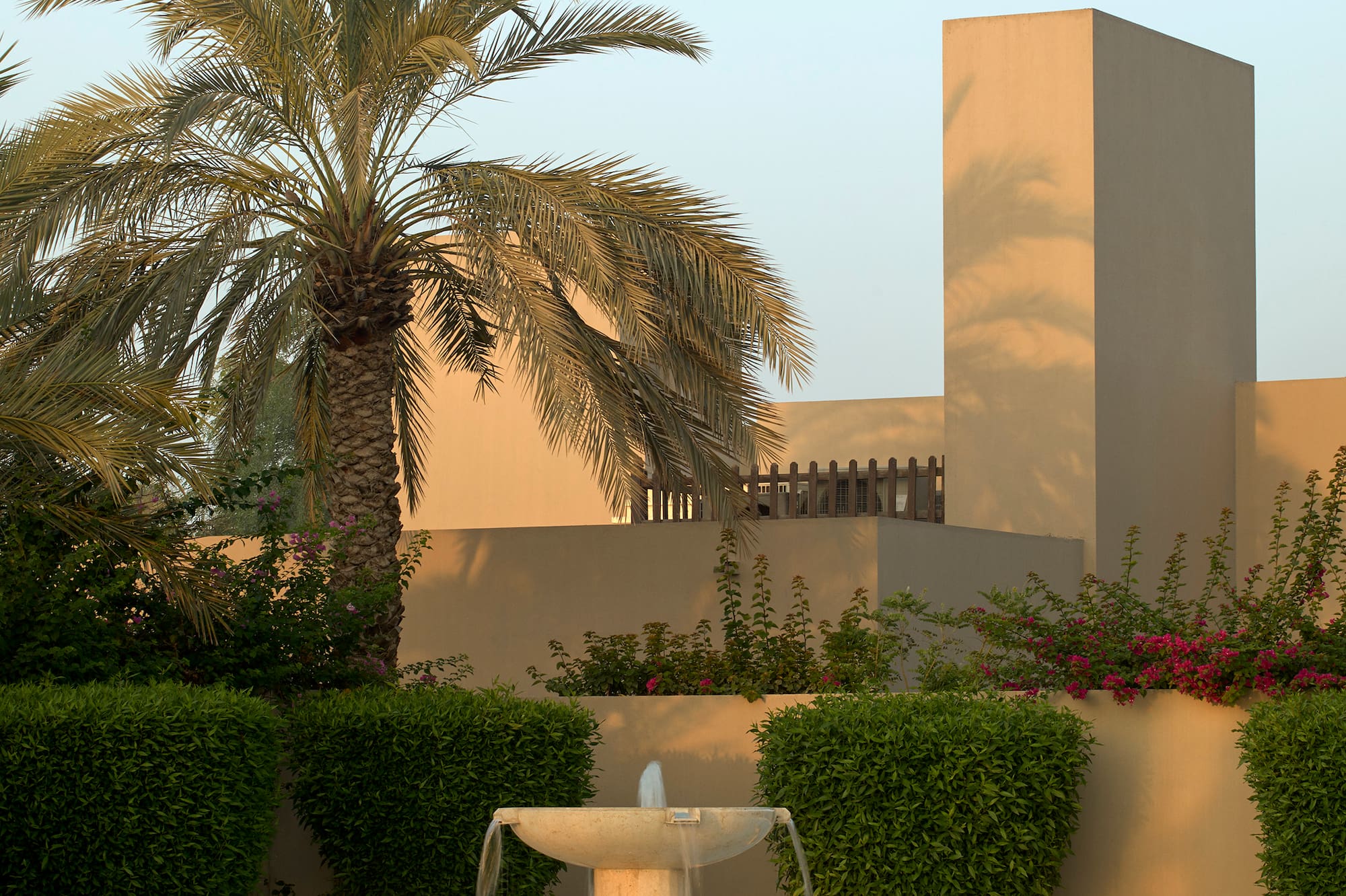 Modern resort garden with stone fountain, palm tree, and tan buildings.