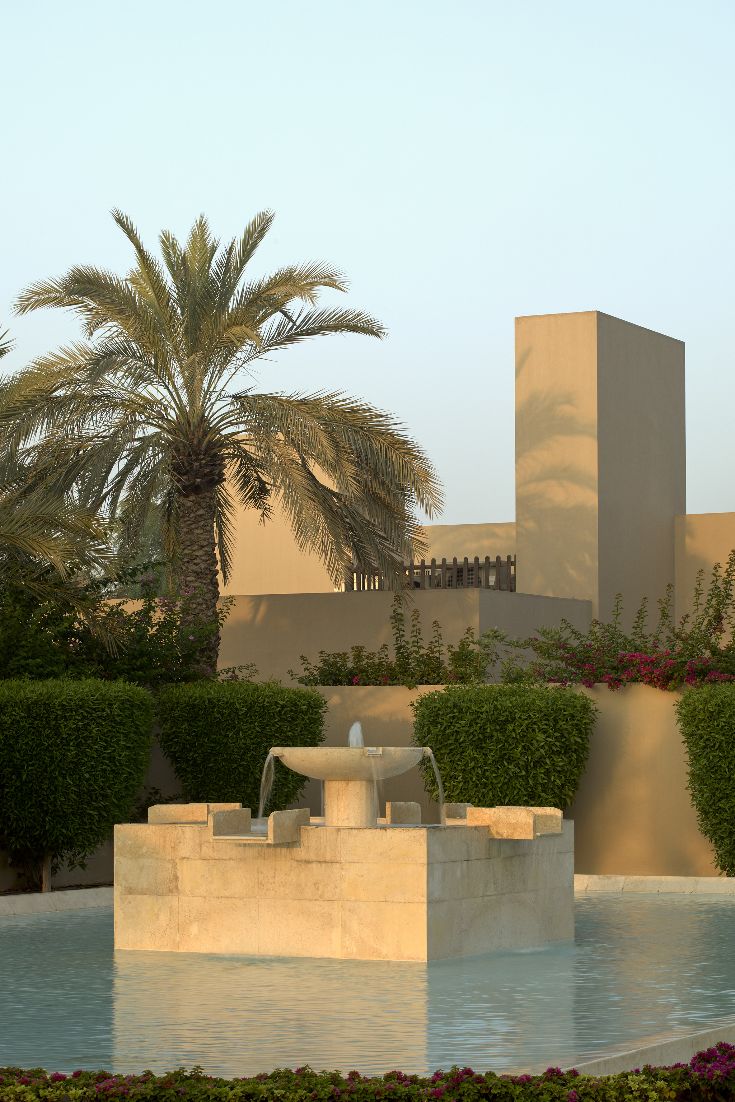 Modern resort garden with stone fountain, palm tree, and tan buildings.
