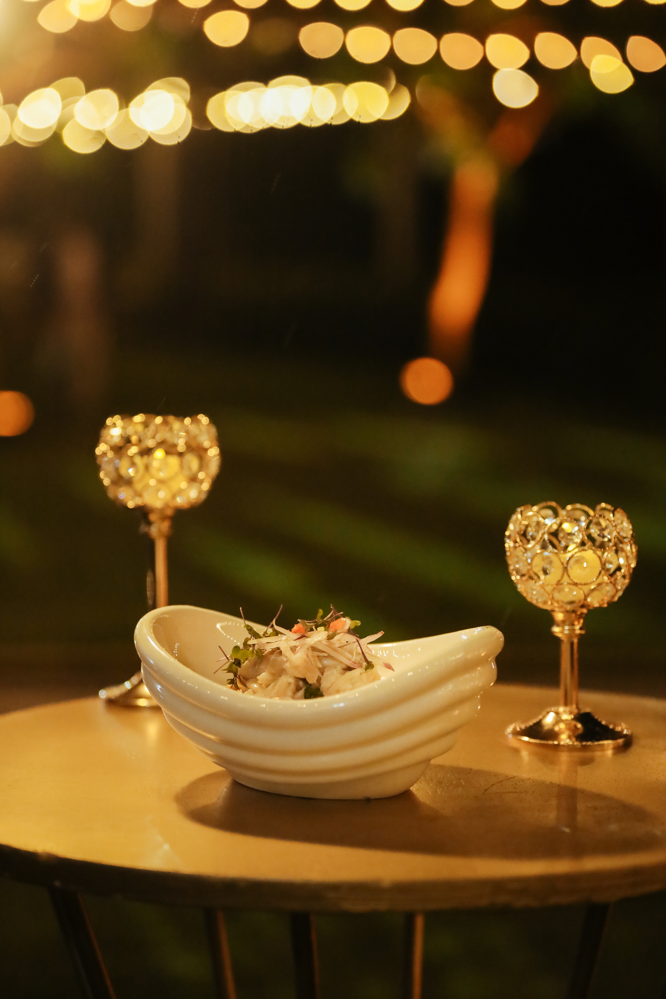 Plato de ceviche en mesa con copas doradas, luces bokeh nocturnas.