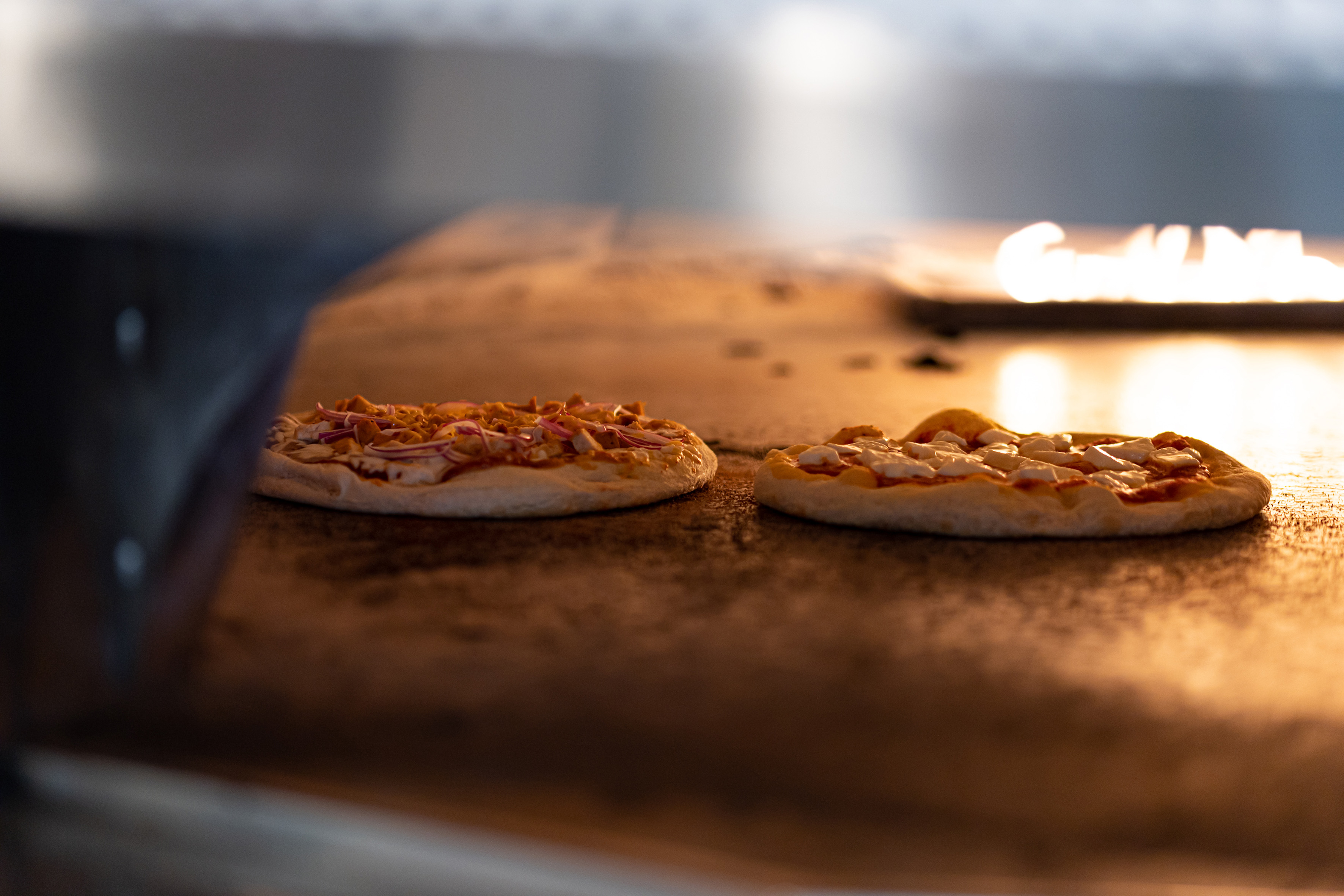 Two fresh pizzas baking in a hot oven with visible flames.