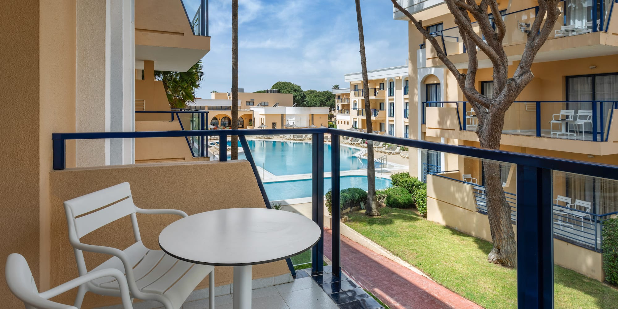 Hotel balcony with table and chairs overlooking a resort pool and palm trees.