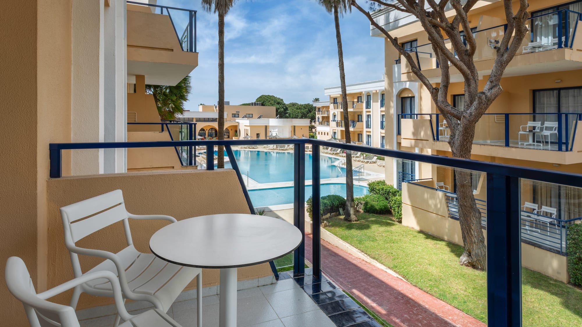 Resort balcony with pool view, white outdoor furniture, and palm trees.