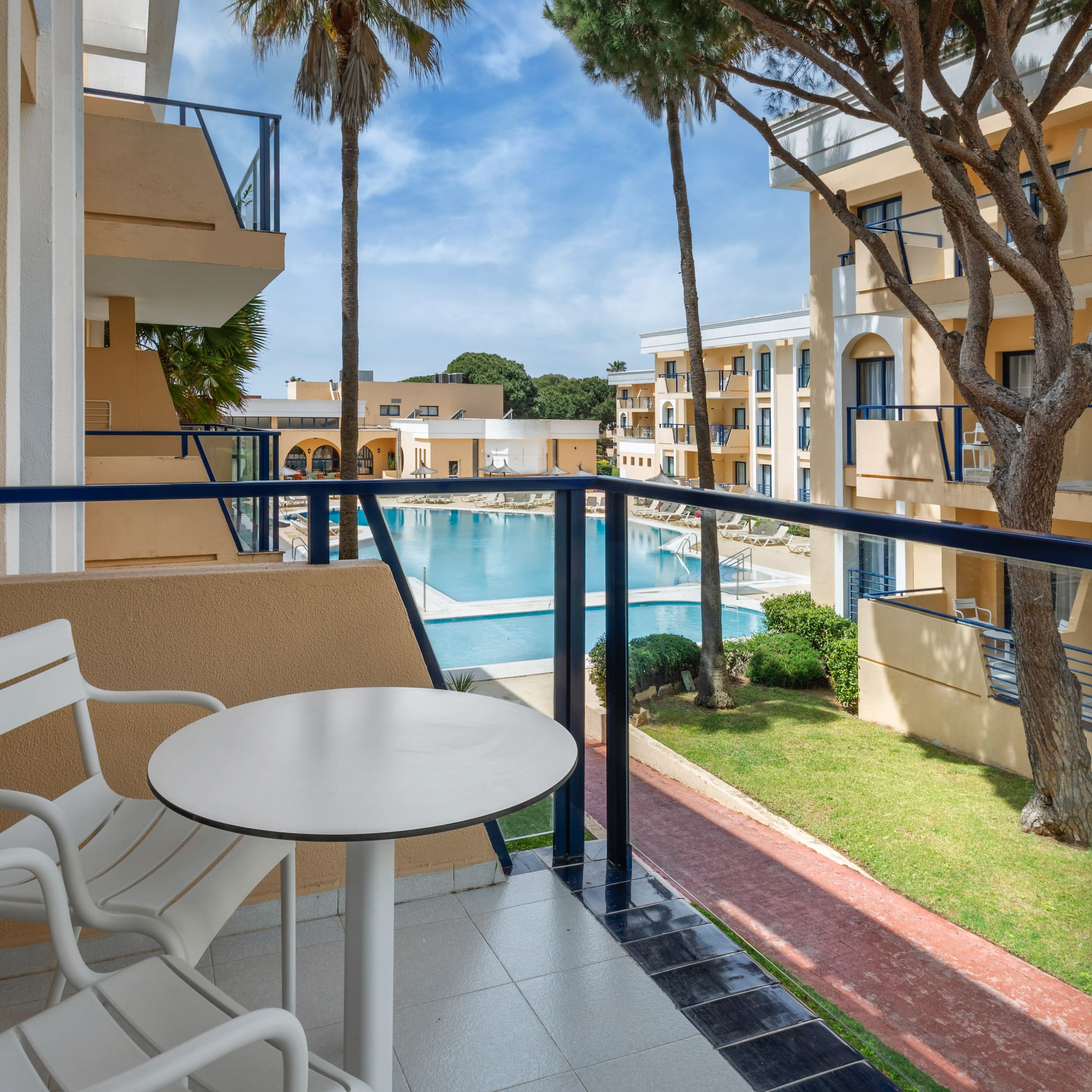 Hotel balcony with table and chairs overlooking a resort pool and palm trees.