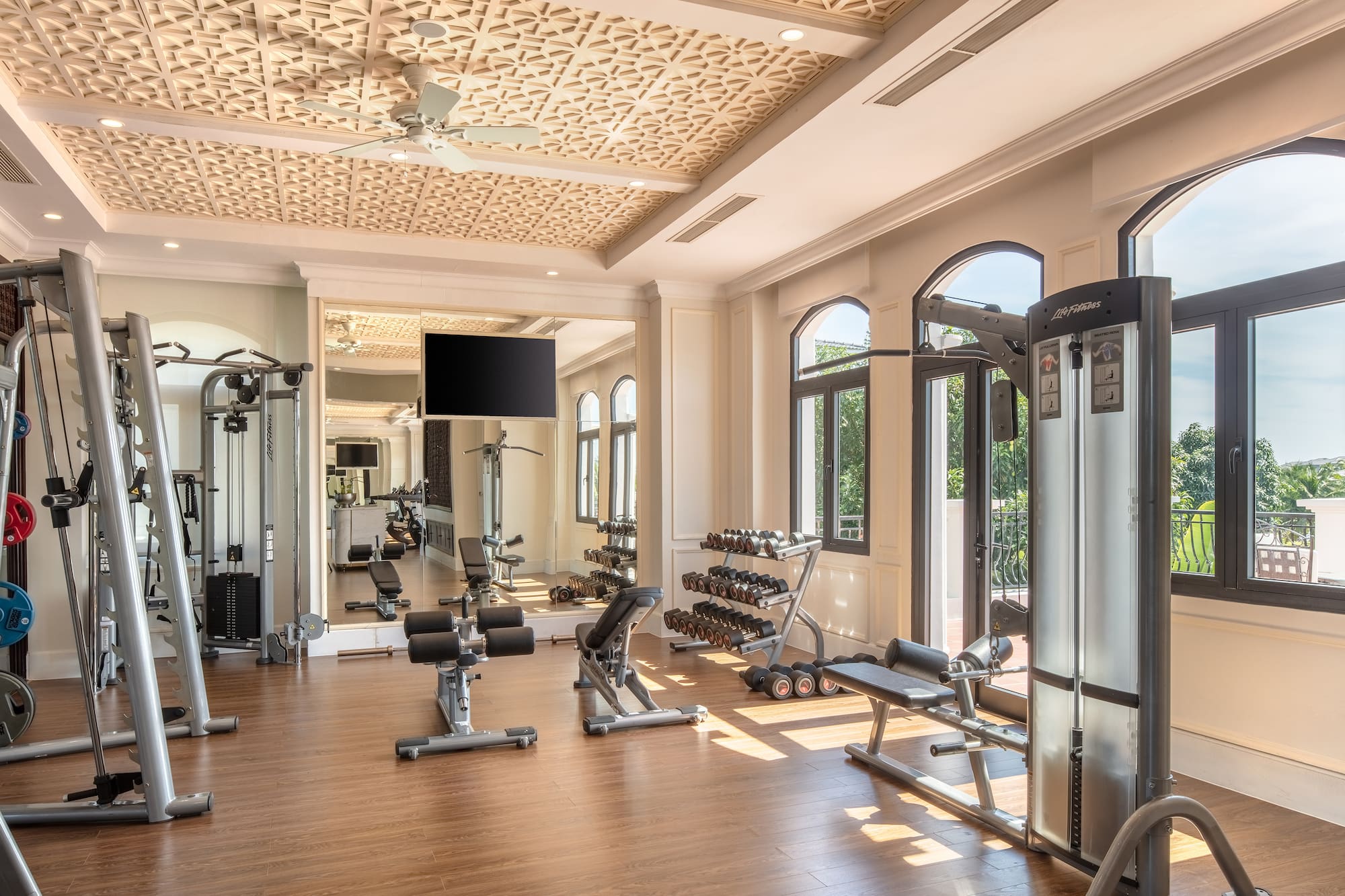 Luxury resort gym with varied exercise equipment, dumbbells, and arched windows.