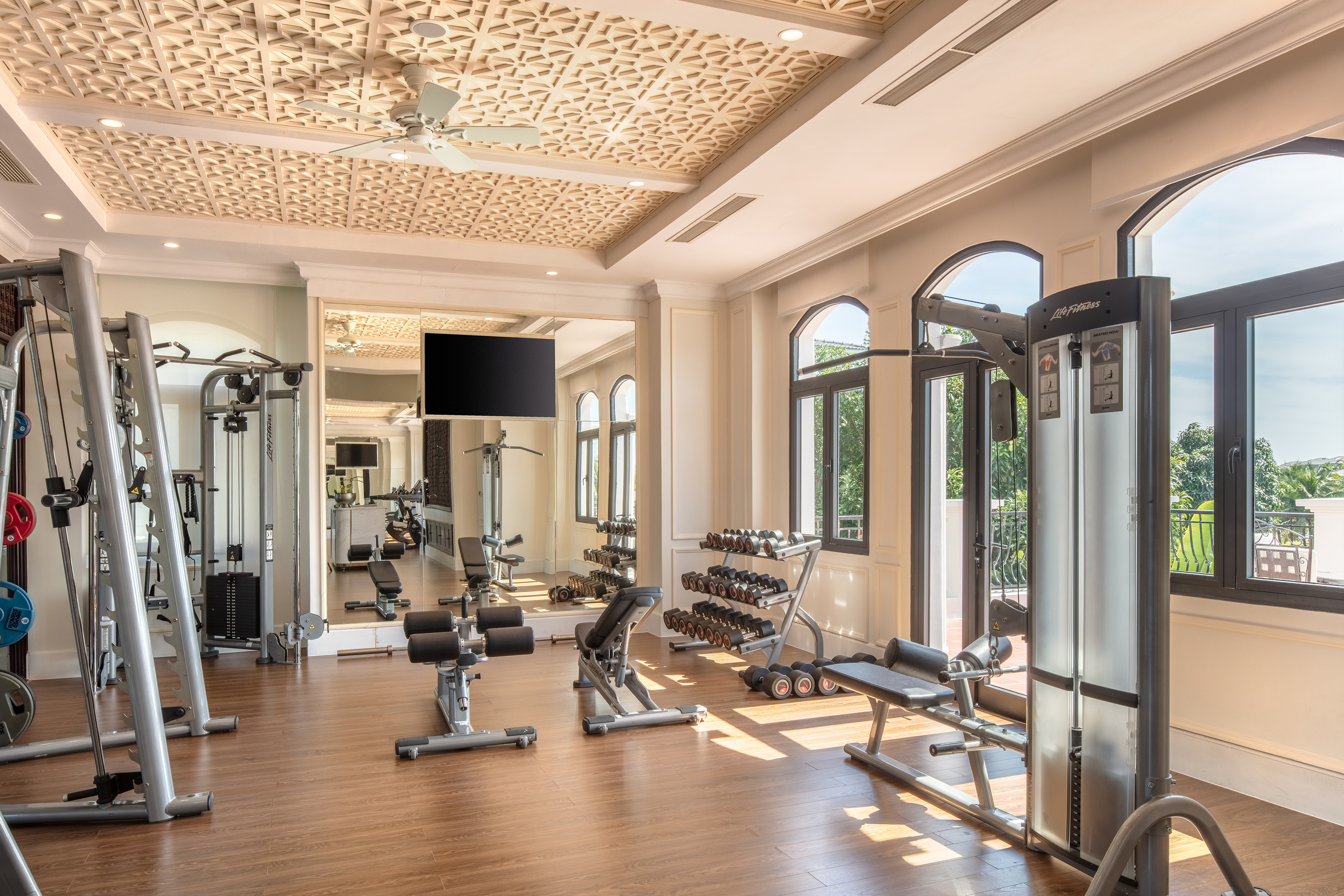 Luxury resort gym with varied exercise equipment, dumbbells, and arched windows.