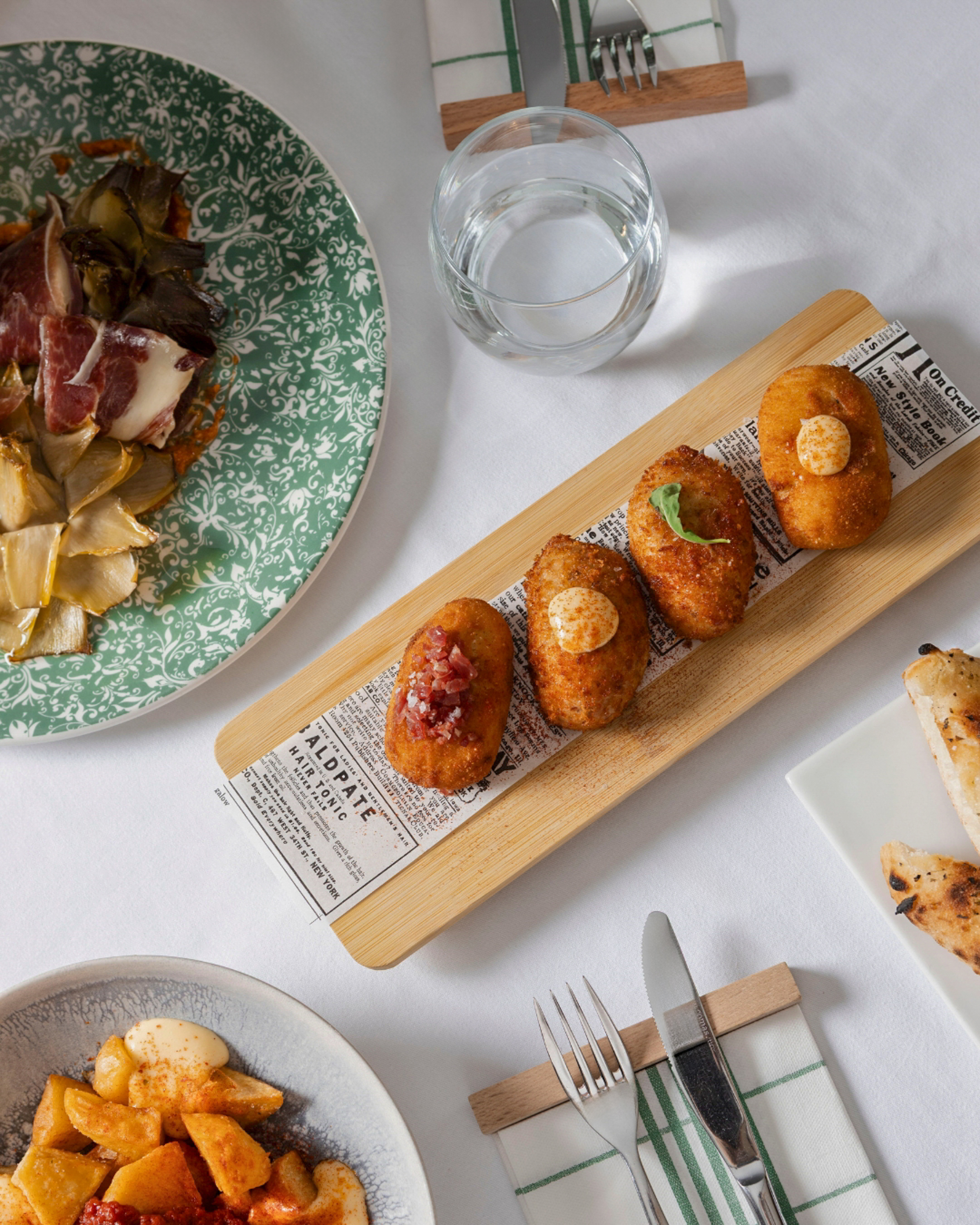 Spanish tapas spread: croquettes, ham, artichokes, and patatas bravas on a table.