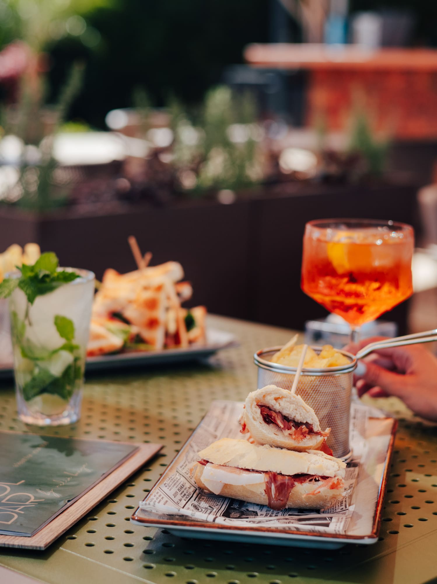 Outdoor patio dining with sandwiches, fries, mojito, and Aperol Spritz.
